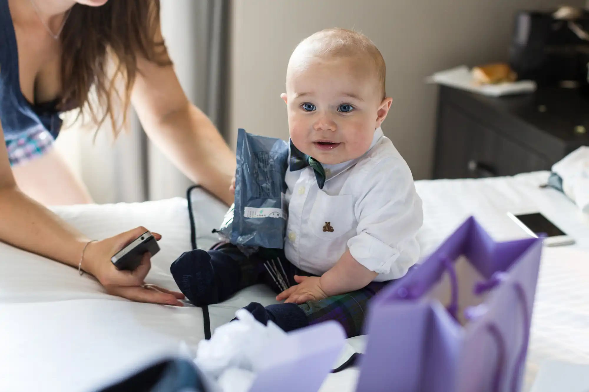 A baby wearing a white shirt, checked trousers, and a bow tie sits on a bed surrounded by gift bags. An adult, partially visible, supports the baby with one hand and holds a mobile with the other. The baby has fair hair and blue eyes and looks slightly to the side.