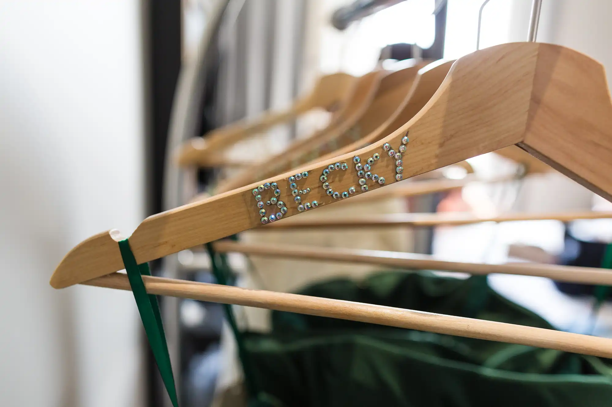 A wooden clothes hanger with “Becky” spelt out in rhinestones is holding a green dress. Other wooden hangers and clothing are visible in the blurred background, with sunlight streaming in from a nearby window.