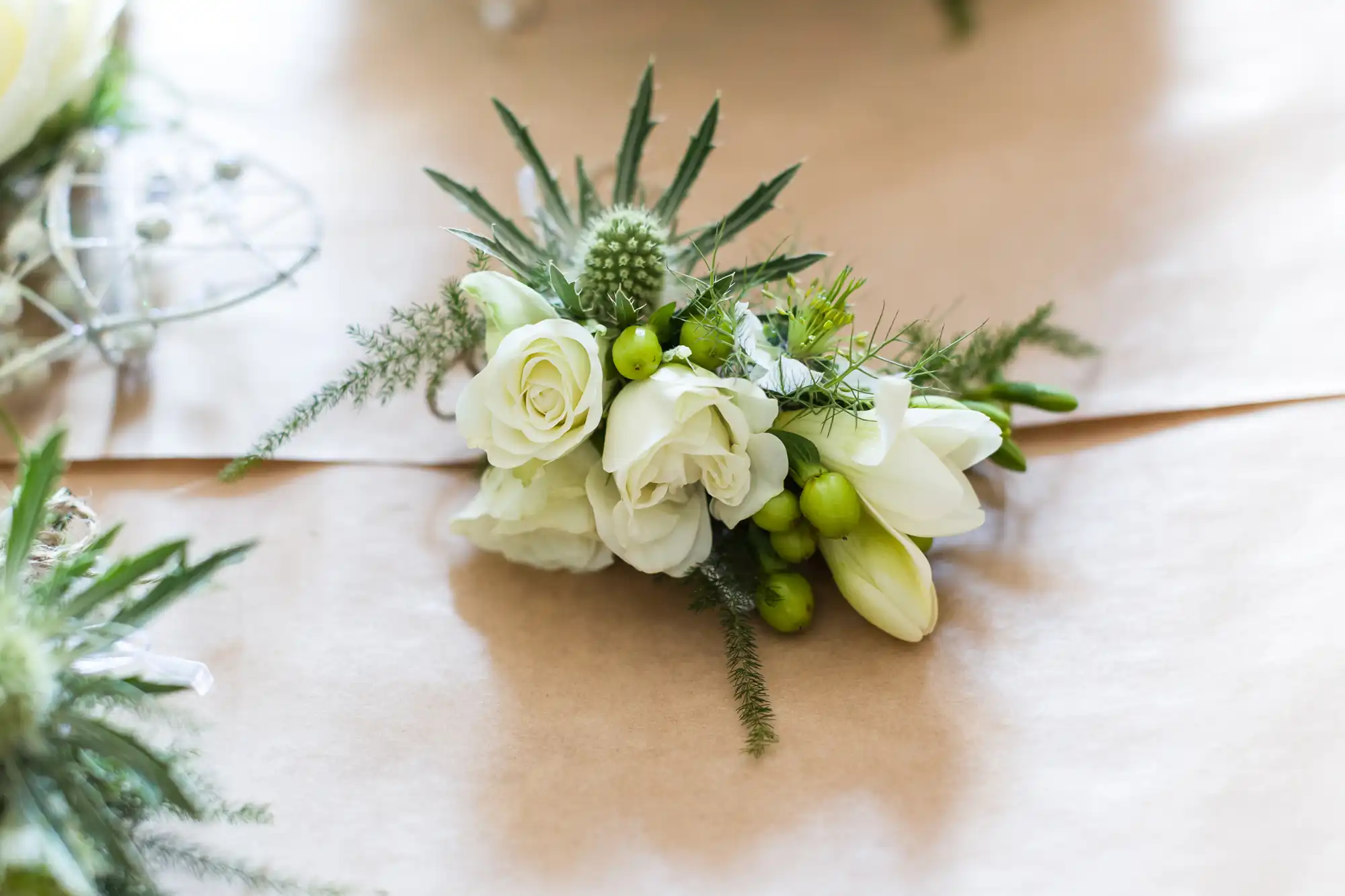 A delicate corsage made of white roses, green berries, and spiky greenery rests on a tan paper surface, with soft natural light highlighting its fresh, elegant arrangement.