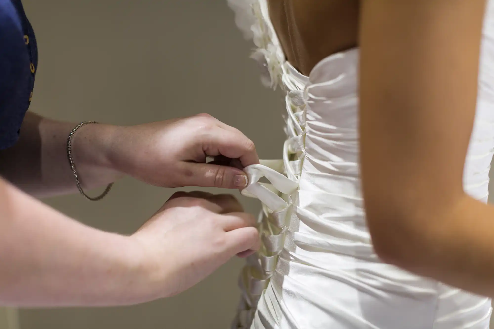 Close-up of hands tying the laces on the back of a white, fitted wedding dress being worn by a bride. The dress features a corset-style lace-up design. The person tying the dress wears a silver bracelet. The background is softly blurred.