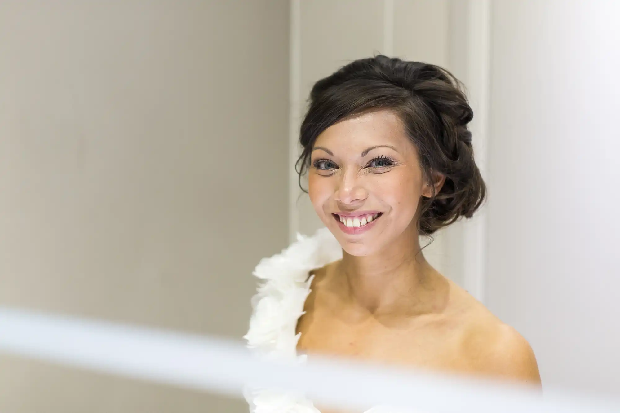 A smiling woman with dark hair styled in an elegant updo wears a white, one-shoulder dress with feathered details. She stands indoors against a softly lit, neutral background, looking directly at the camera.