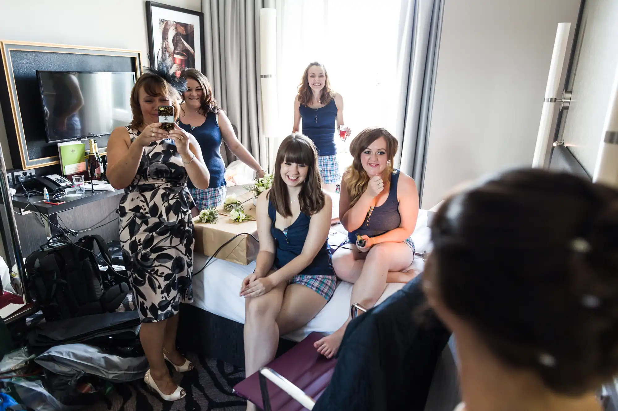 Five women are in a bright, cluttered hotel room. Three sit on a bed in matching blue vests and checked shorts, smiling. One stands behind the bed and another woman in a floral dress takes a photo. A woman’s head is visible in the foreground.