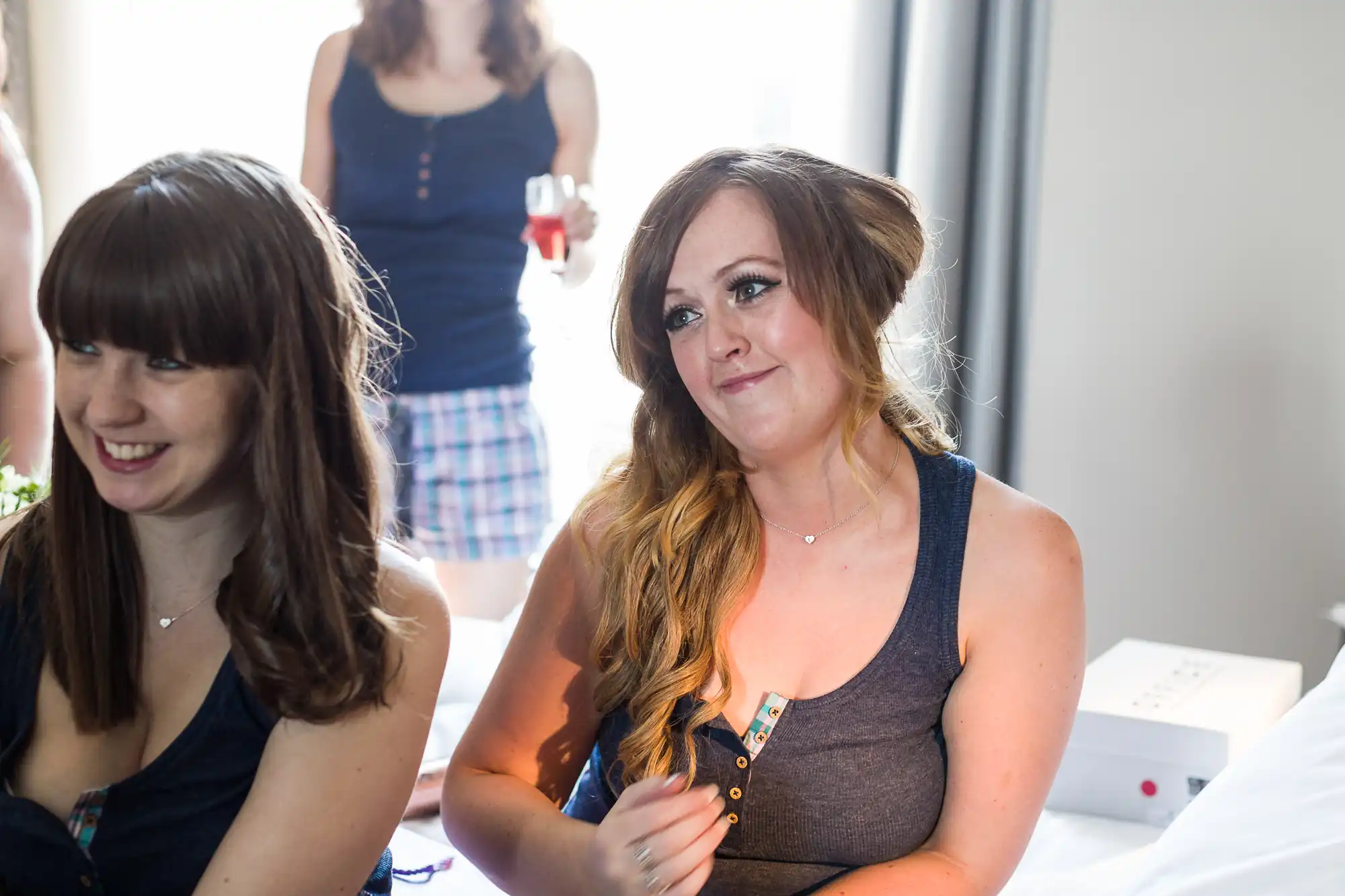 Two women sit indoors on a bed, wearing navy vests. The woman on the right has long, wavy hair and smiles softly, while the woman on the left with straight hair laughs. A woman in the background holds a drink and wears a checked skirt and navy top.