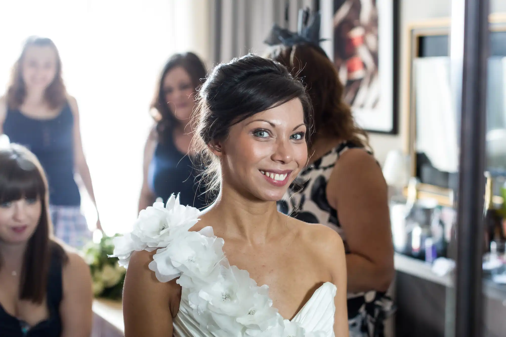 A smiling woman in a white, one-shoulder dress decorated with large floral accents stands indoors. She has dark hair in an elegant up-do. Several other women, slightly out of focus, are in the background in a well-lit room with artwork and a mirror.
