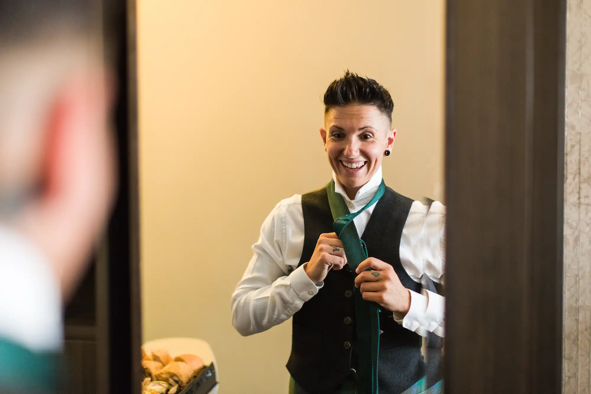 A person with short dark hair and wearing a waistcoat over a white shirt smiles in a mirror while tying a green tie. The background is softly lit, with some objects blurred on a counter, creating a warm, candid atmosphere.