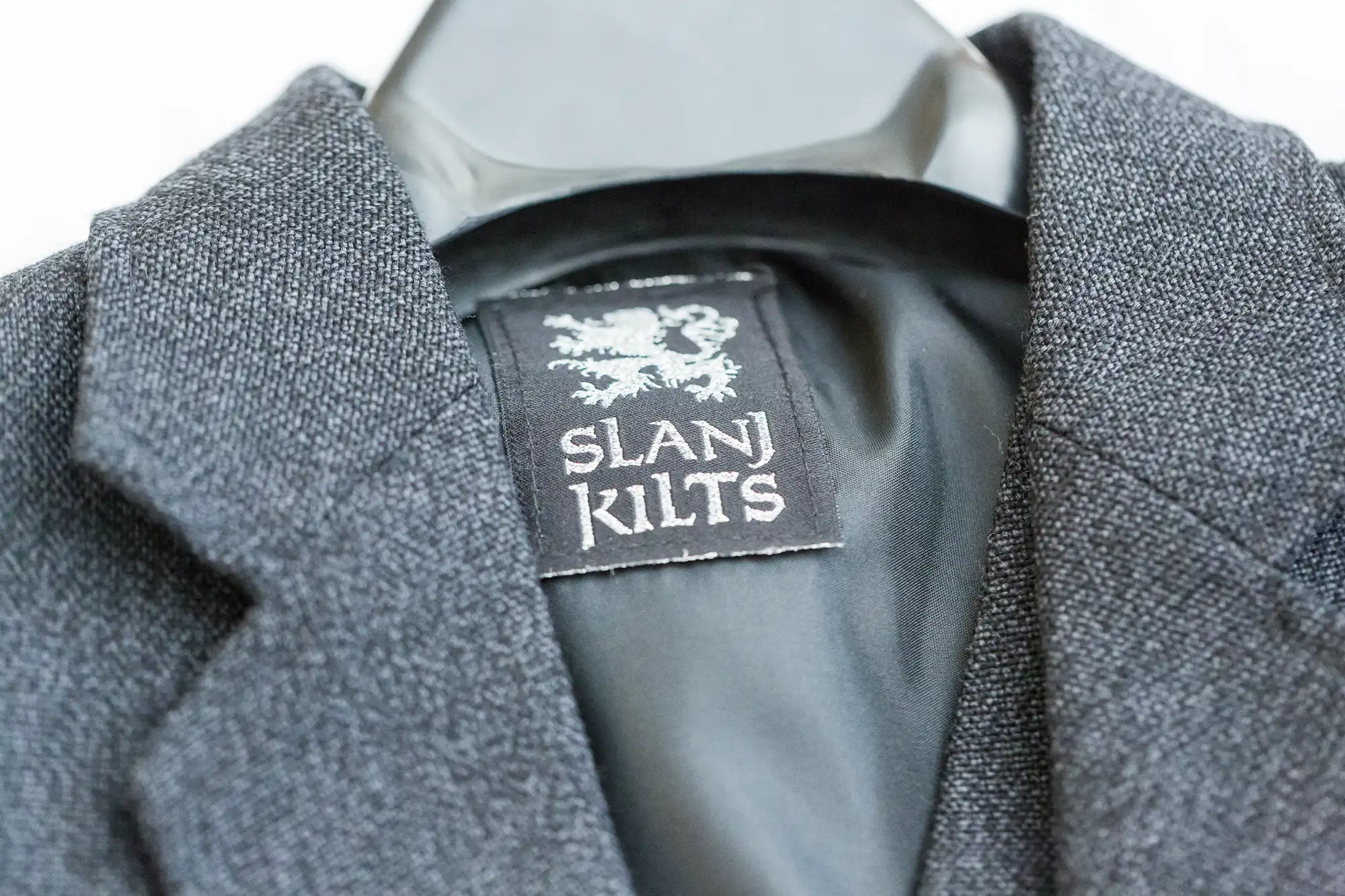 Close-up of a dark grey, textured blazer on a black hanger. The label inside the collar reads "SLANJ KILTS" in white text, with a lion graphic above the text. The inner lining of the blazer is smooth and black.