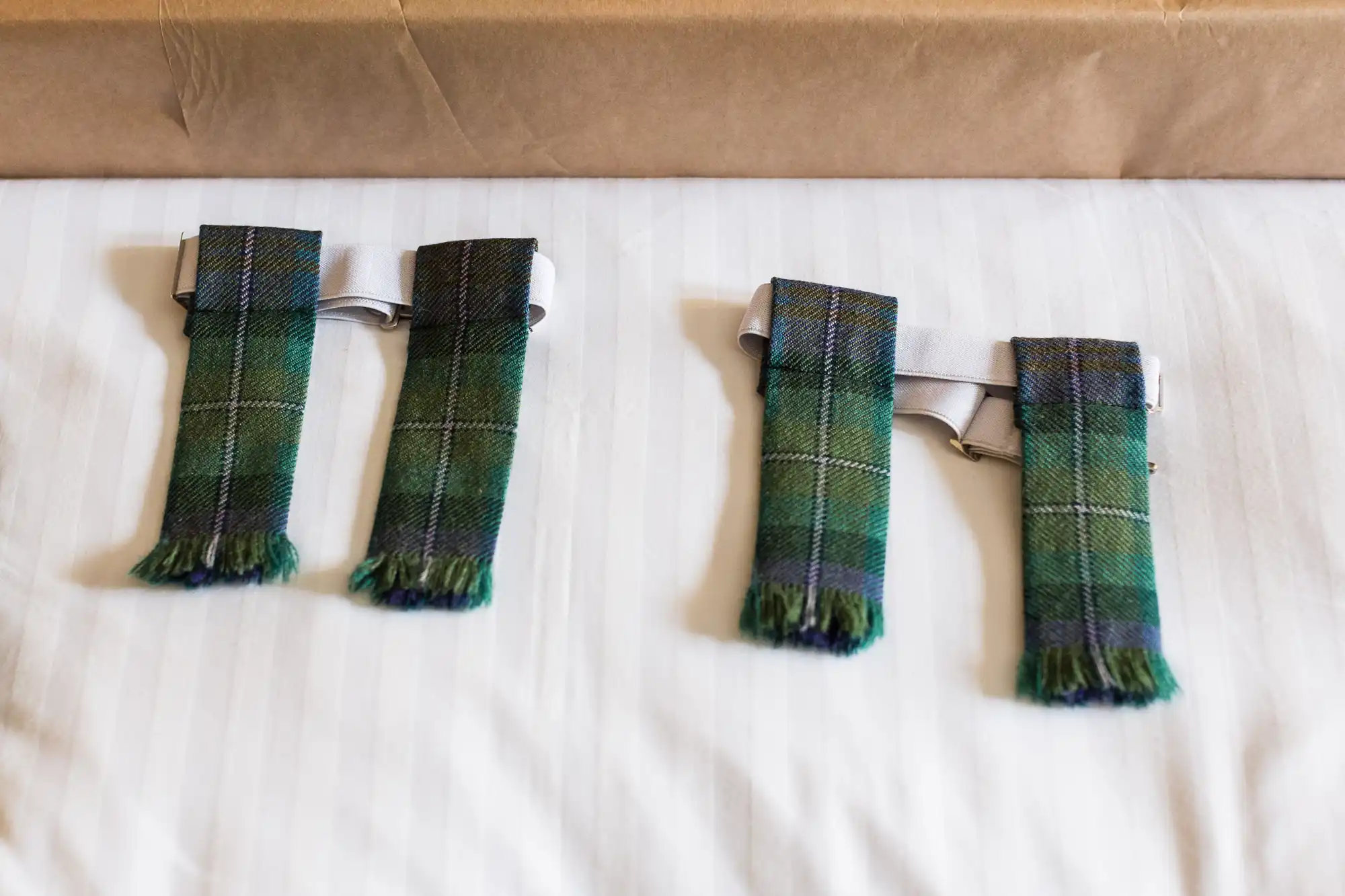 Two pairs of green tartan kilt flashes with blue, black, and yellow stripes lie side by side on a white, striped fabric surface. The flashes have frayed ends and are attached to silver garters. A brown box is partially visible at the top of the image.
