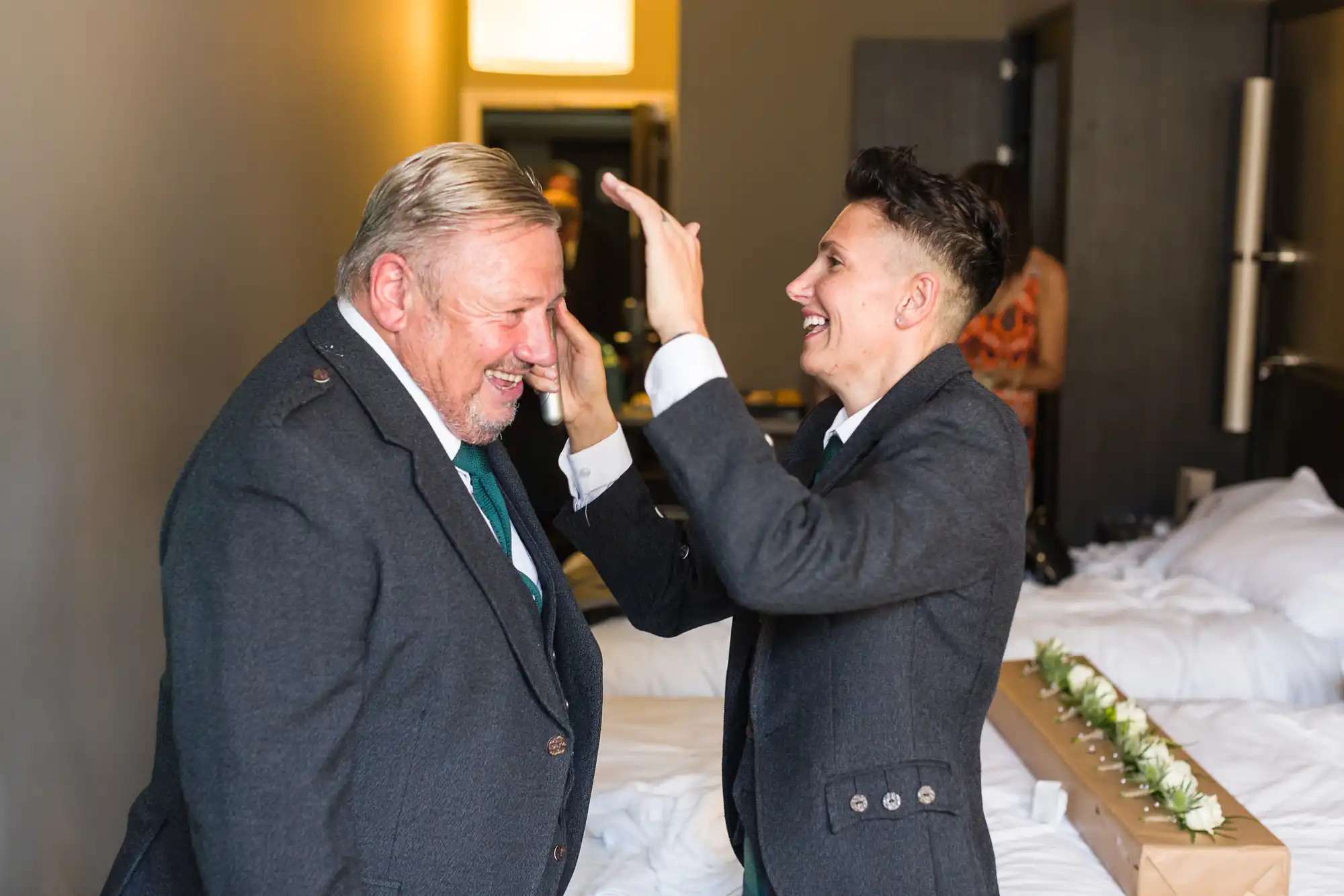 Two people in suits share a joyful moment in a hotel room; one, older with grey hair, smiles as the younger, with short dark hair, playfully fixes his appearance. Beds with white linen and a flower garland are visible in the background.