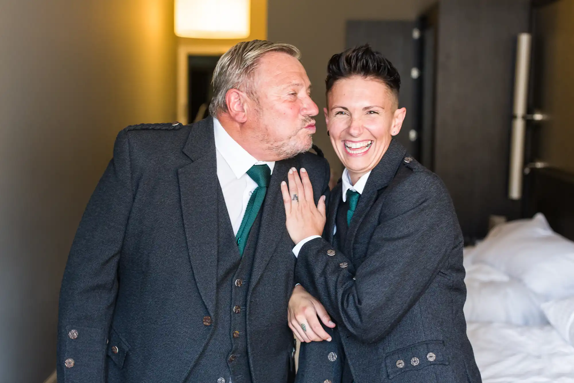 Two people in matching grey suits and green ties smile joyfully in a well-lit room. One person leans in to kiss the other's cheek, while the other laughs with their hand on their partner's chest, showing a wedding ring. A bed and lamp are visible in the background.