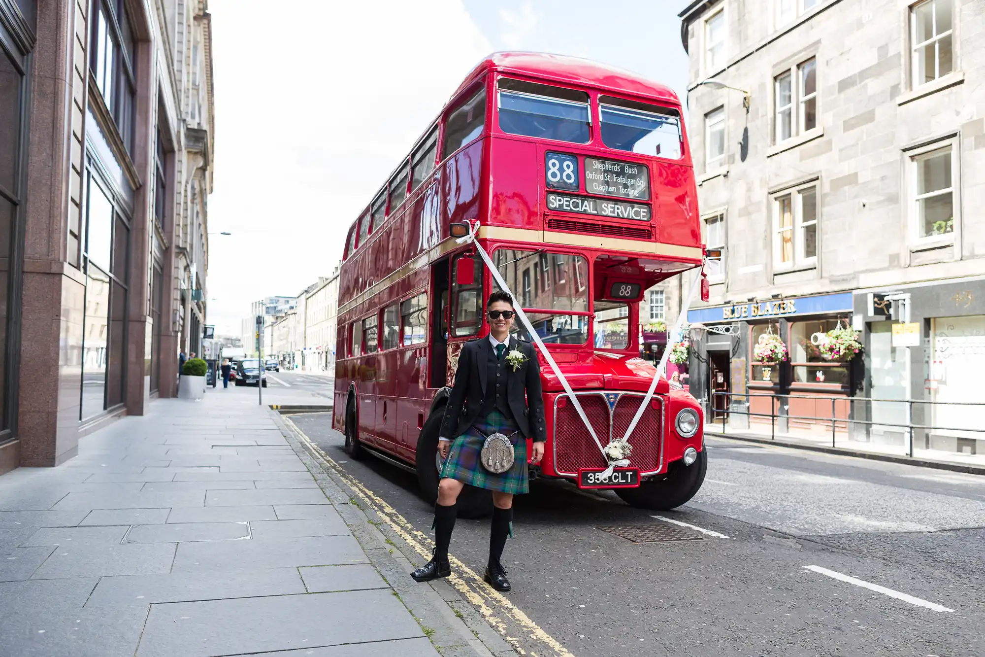 A person in a traditional Scottish kilt outfit stands smiling in front of a classic red double-decker bus on a city street. The bus displays route 88 and "Special Service." Stone buildings line the street on a partly cloudy day.