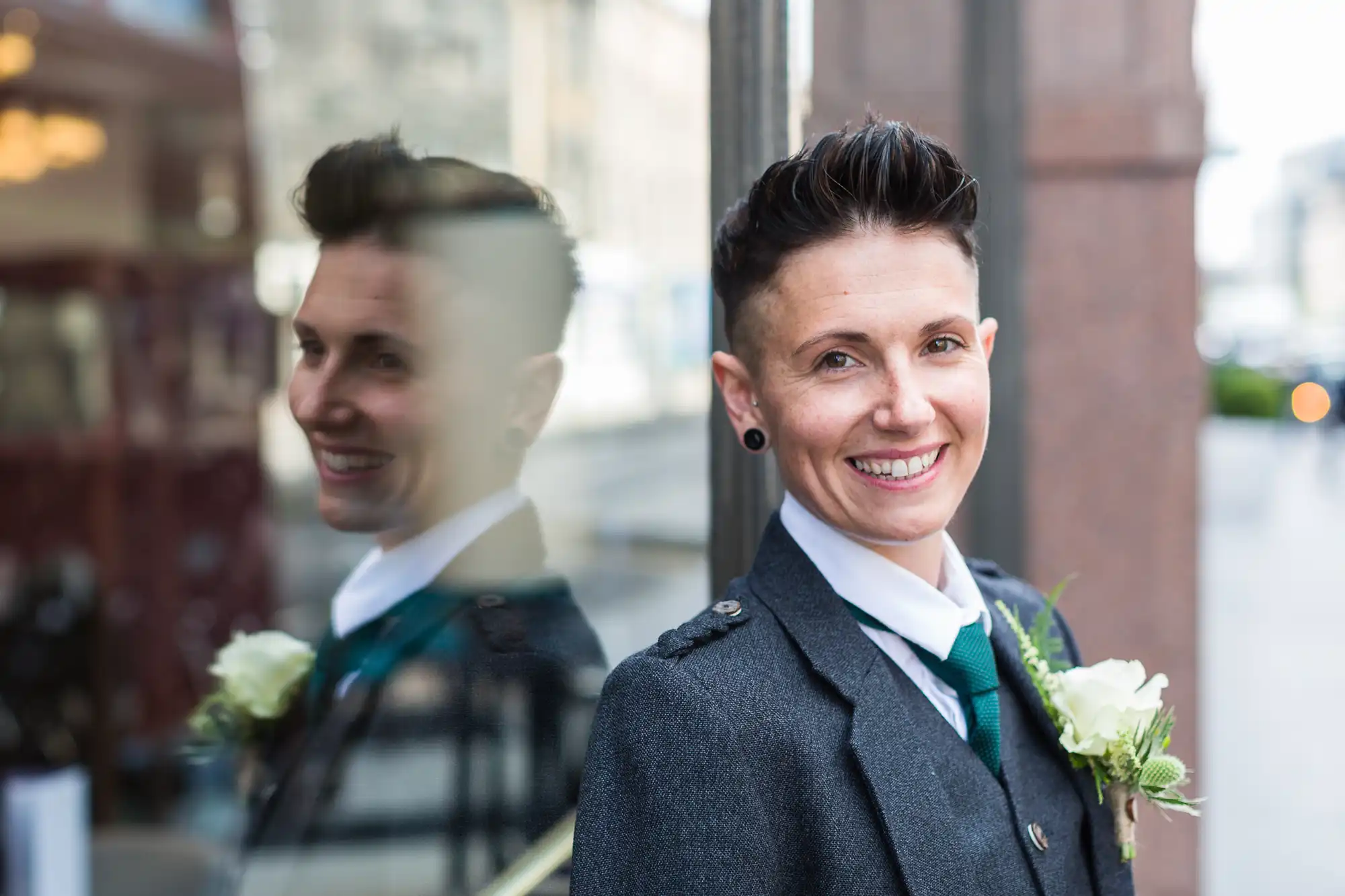 A person with short dark hair, wearing a grey suit jacket, white shirt, and green tie, smiles whilst standing next to a reflective glass wall. They have a white rose buttonhole and round black earrings. Their reflection is visible in the glass.
