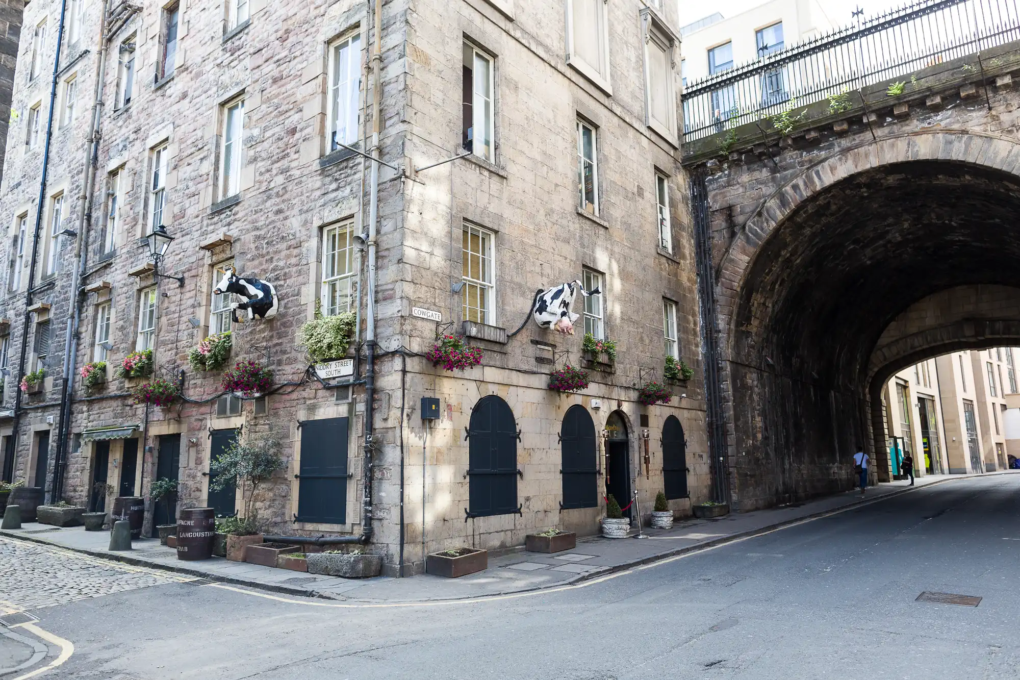 A stone building with black shutters and flower boxes has two lifelike cow statues projecting from its wall. An arched stone bridge covers part of the street beside the building. The scene appears quiet and empty.