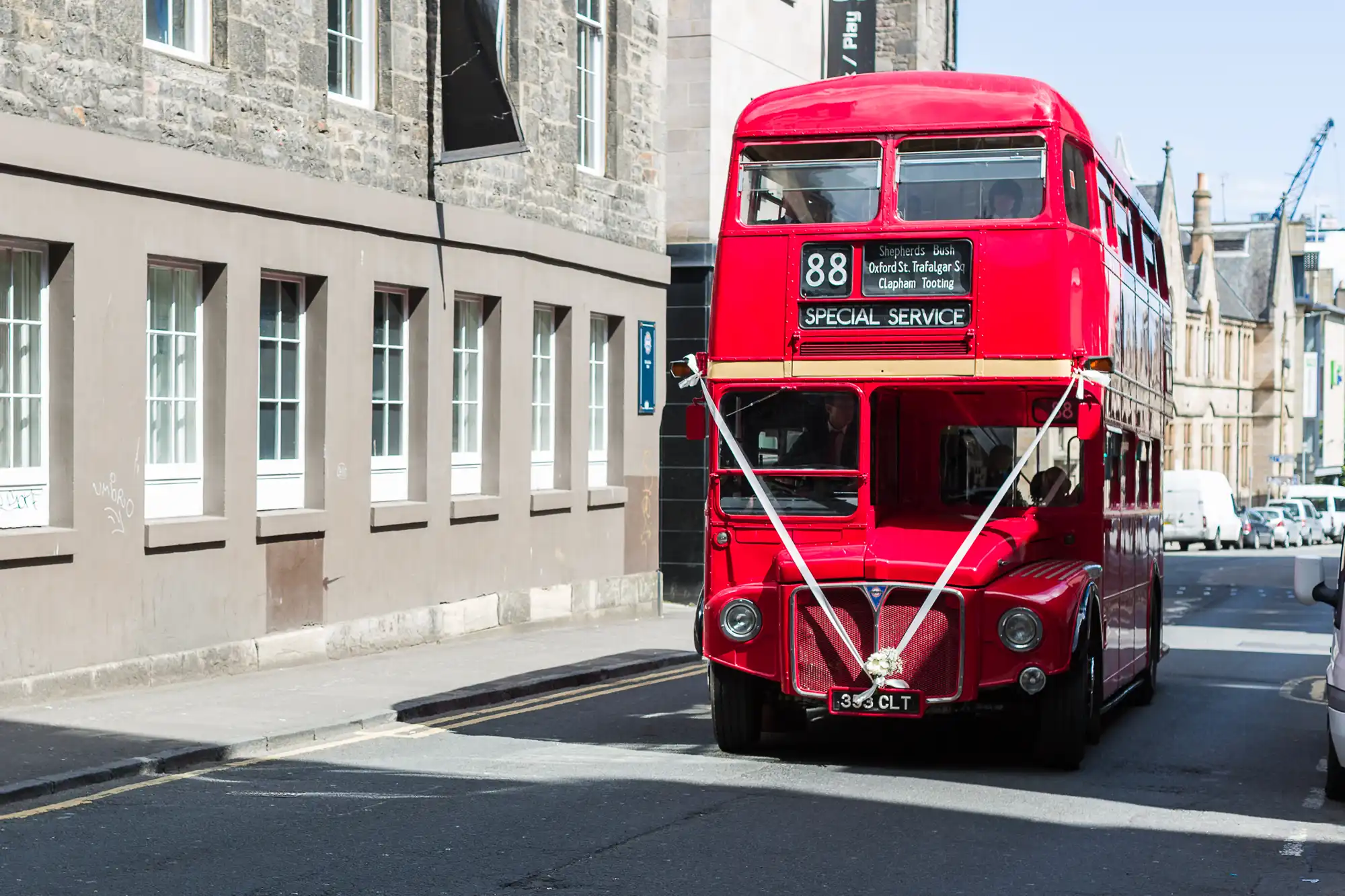A classic red double-decker bus decorated with white ribbons is parked on a sunny city street. The sign reads "88 Special Service" and the route includes "Muirhouse, Granton." Old stone buildings line the background.