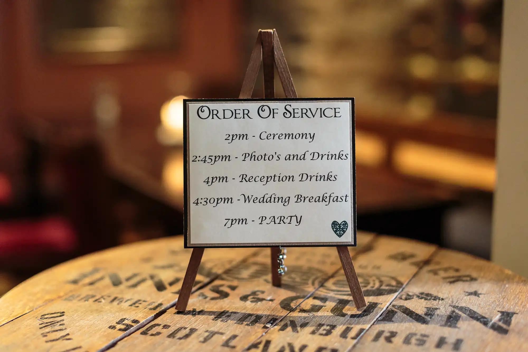 A small wooden easel on a barrel displays a wedding order of service sign listing times for the ceremony, drinks, photos, reception, wedding breakfast, and party, with elegant script and a small heart in the bottom right corner. Background is softly blurred.