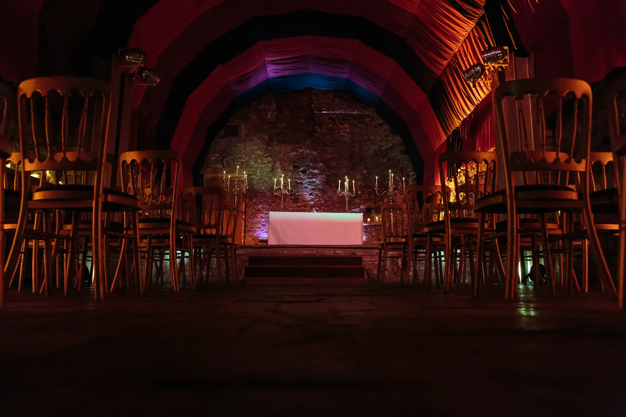 Rows of empty golden chairs face a white-draped table with candelabra, set on a small stage against a stone wall. The arched ceiling is draped in red and purple fabric, creating a dramatic, dimly lit atmosphere reminiscent of a ceremonial or event space.