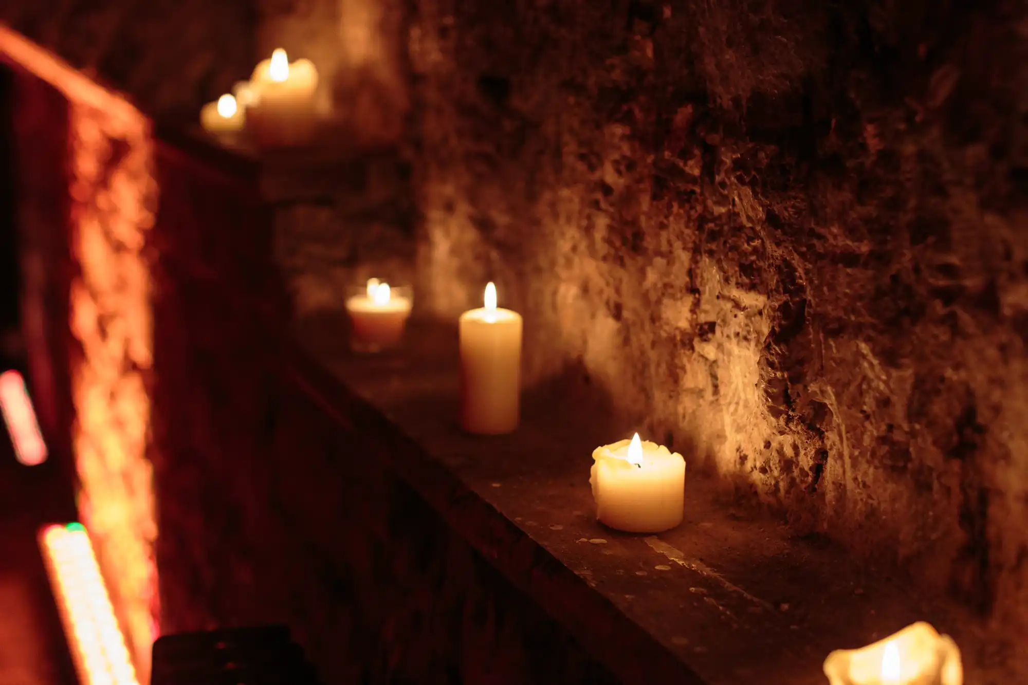 Several lit candles are placed in a row on a rough, stone ledge. The warm candlelight softly illuminates the textured stone wall, creating a cosy, intimate atmosphere in a dimly lit space.