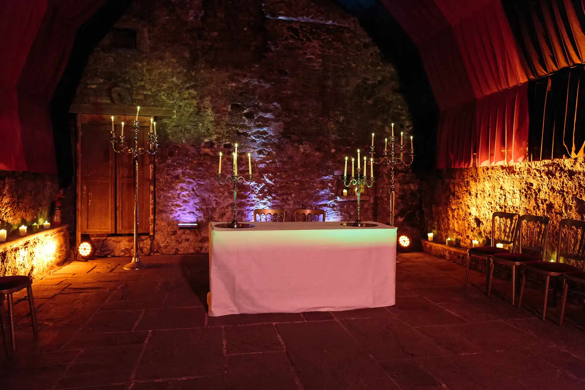 A dimly lit stone room with a white-clothed table in the centre, two plates on top. Ornate candlesticks with lit candles flank the table. Wooden chairs line the walls, and the scene glows with purple and yellow lights.