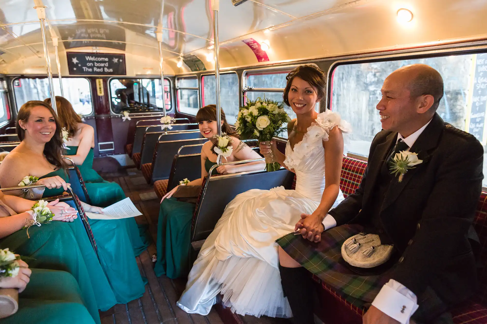 A bride in a white dress sits smiling beside a man in a kilt on a vintage red bus. Bridesmaids in green dresses sit nearby, holding bouquets, all appearing happy. The bus has red tartan seats and a sign that says, “Welcome on board The Red Bus.”.