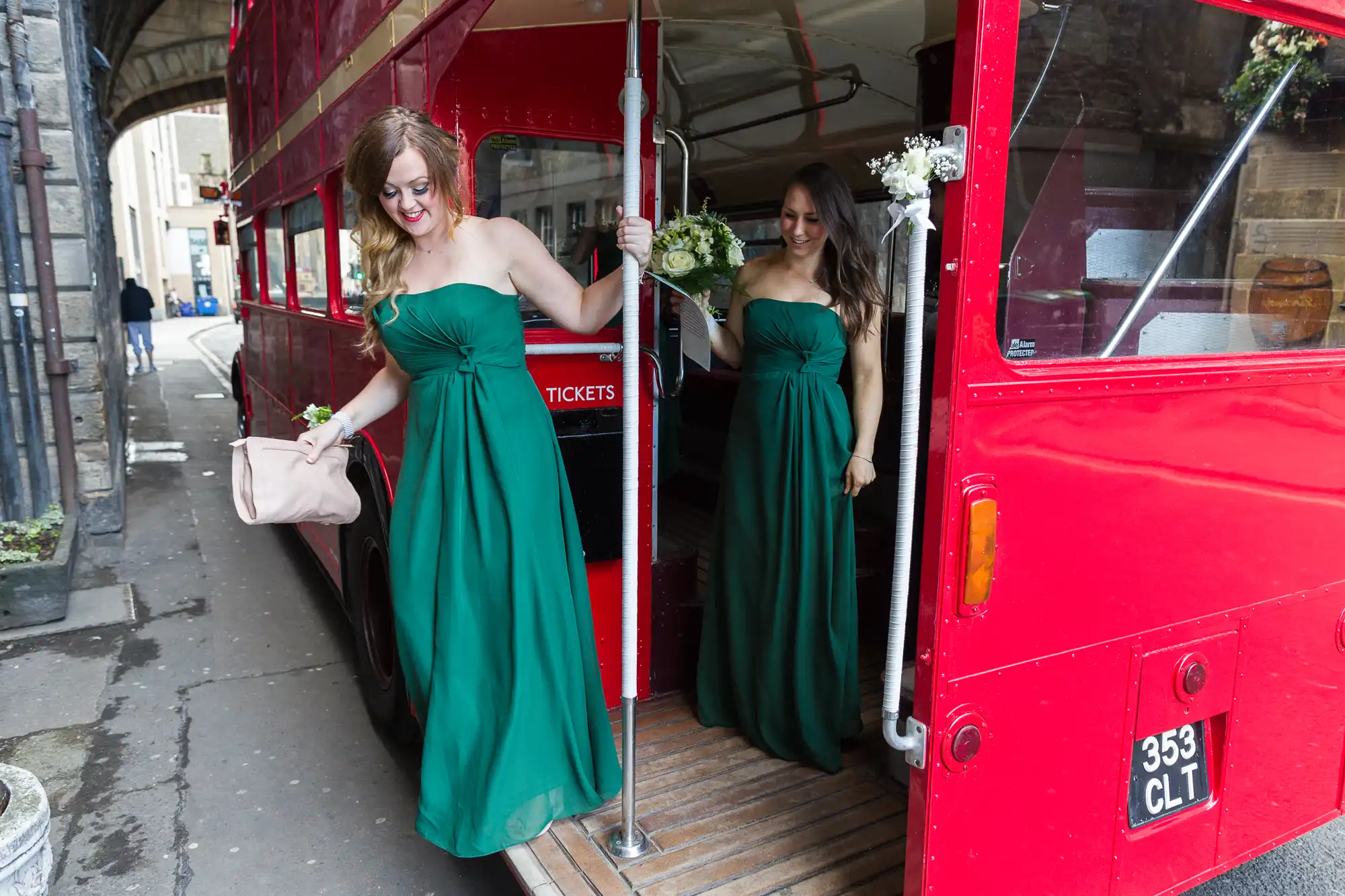 Two women in strapless green dresses step off a vintage red double-decker bus. One is holding a blush pink clutch and smiling, whilst the other holds a bouquet. The bus door is open, showing the wooden floor and floral decorations.