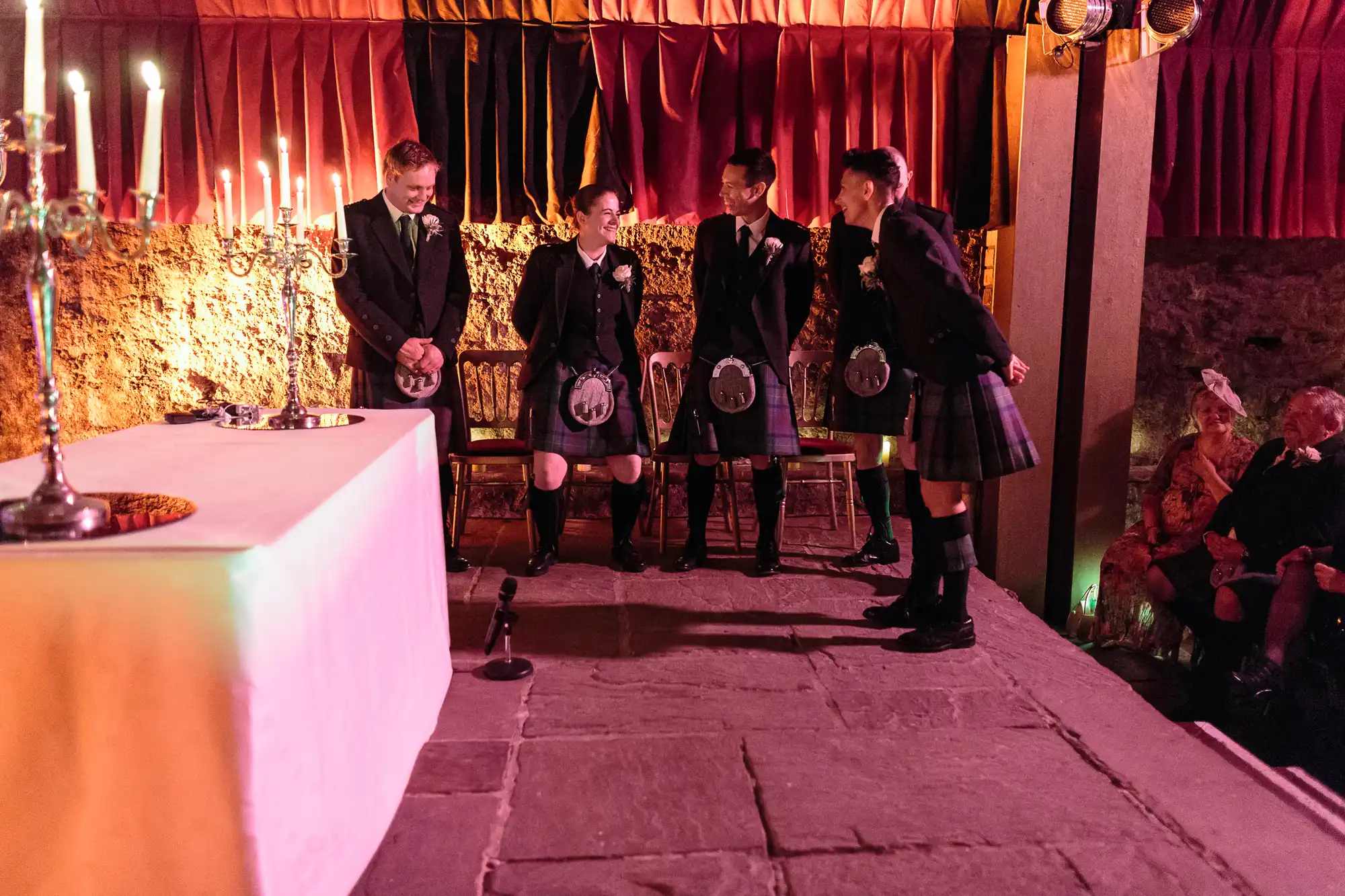 Four people in kilts and jackets stand laughing on a stone stage near a white-clothed table with candles. Behind them are red curtains and seated guests, some dressed formally. Warm lighting gives the scene a festive, celebratory atmosphere.