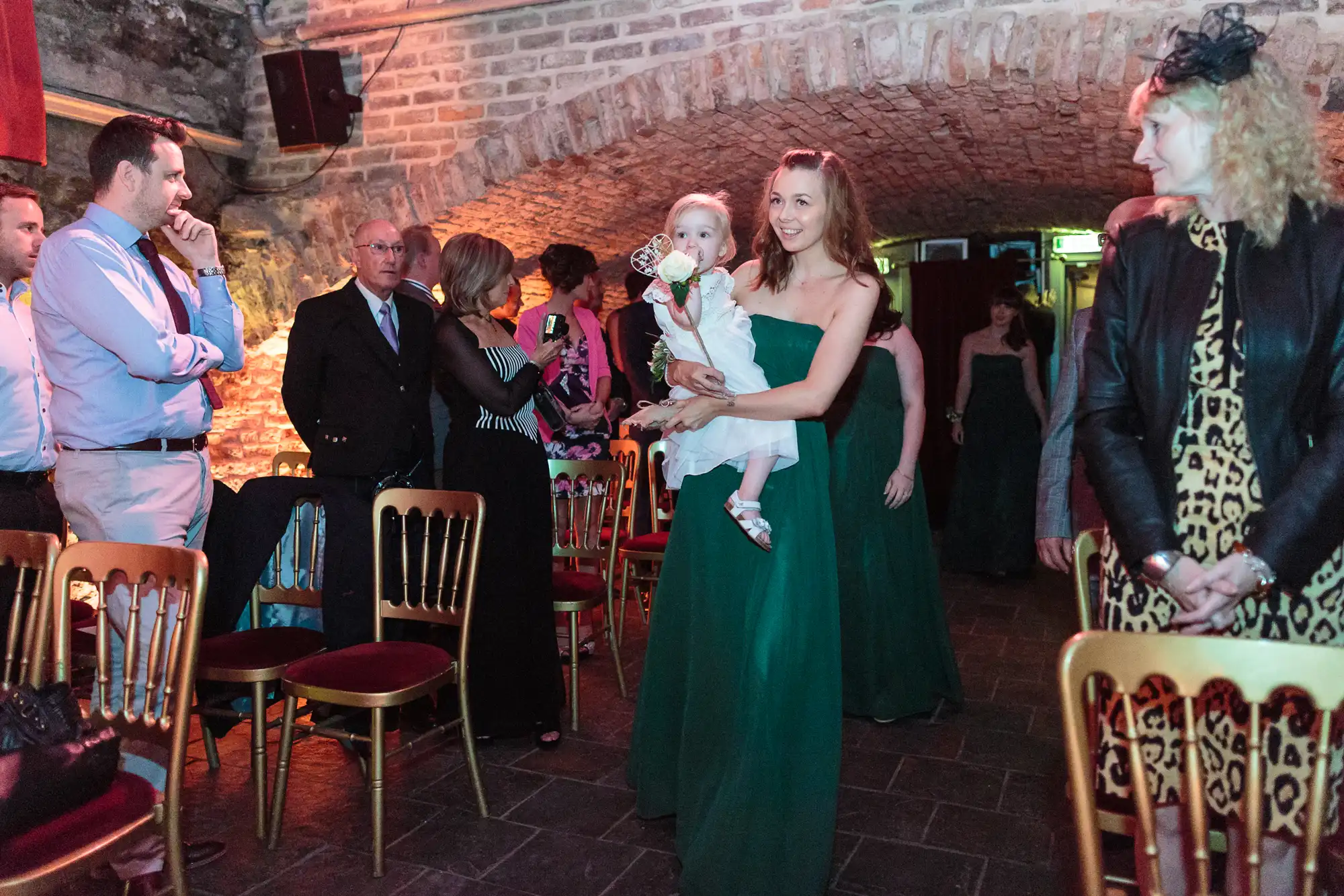 A smiling woman in a green dress walks down an aisle holding a small child. People in formal attire watch her, sitting and standing among red-cushioned chairs, in a brick-vaulted room with warm lighting.