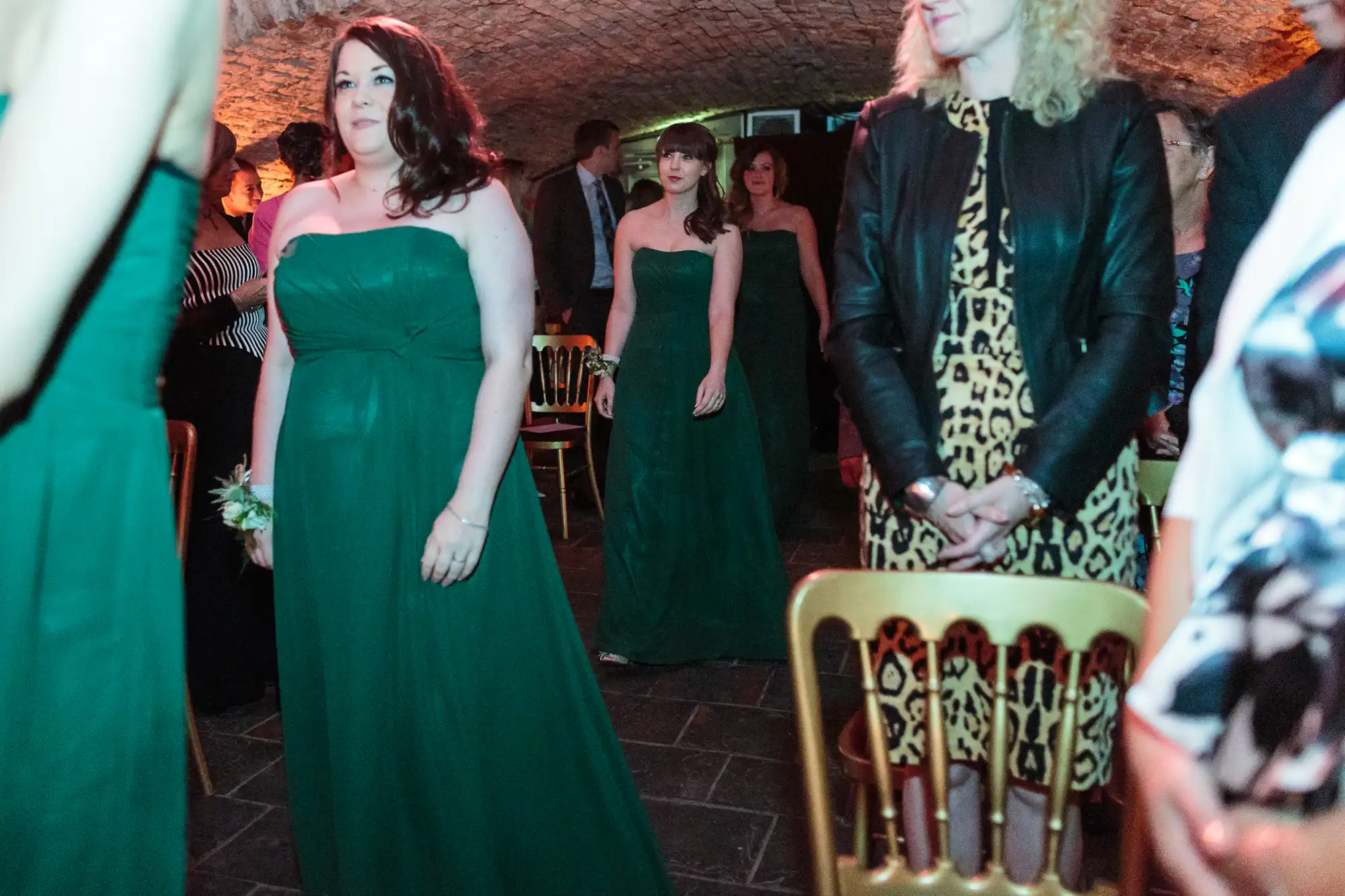 Three women in strapless dark green dresses walk down an aisle at an indoor event with low arched stone ceilings. Guests, including a woman in a leopard print dress and black jacket, are seated on gold chairs along the aisle.