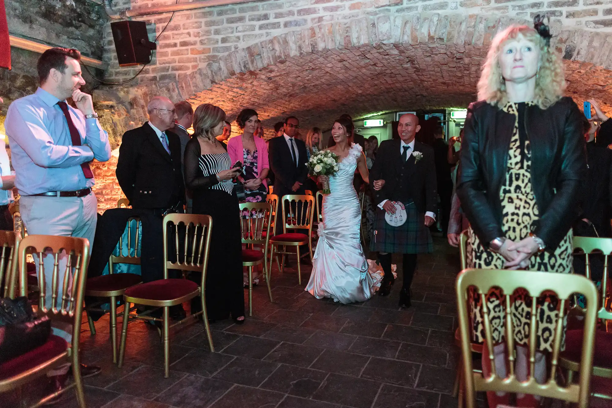 A bride in a white dress walks down the aisle with a man in a kilt, surrounded by standing guests in formal attire. They are in a stone-walled, arched room with red-cushioned chairs. A woman in a leopard print dress stands in the foreground.