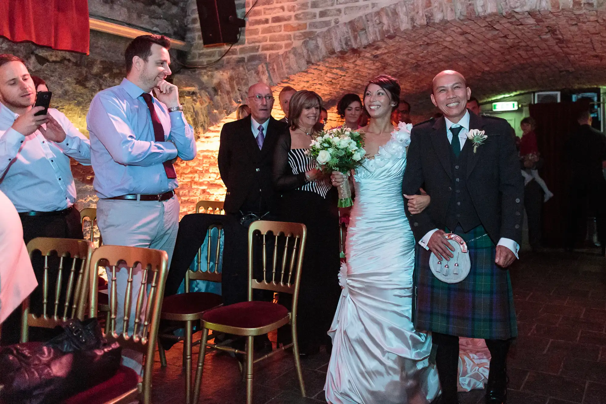 A smiling bride in a white dress and a man in a kilt walk down an aisle in a rustic, brick-walled venue. Guests stand and watch, some taking photos, as they pass by rows of gold chairs. Warm, festive lighting creates a celebratory atmosphere.