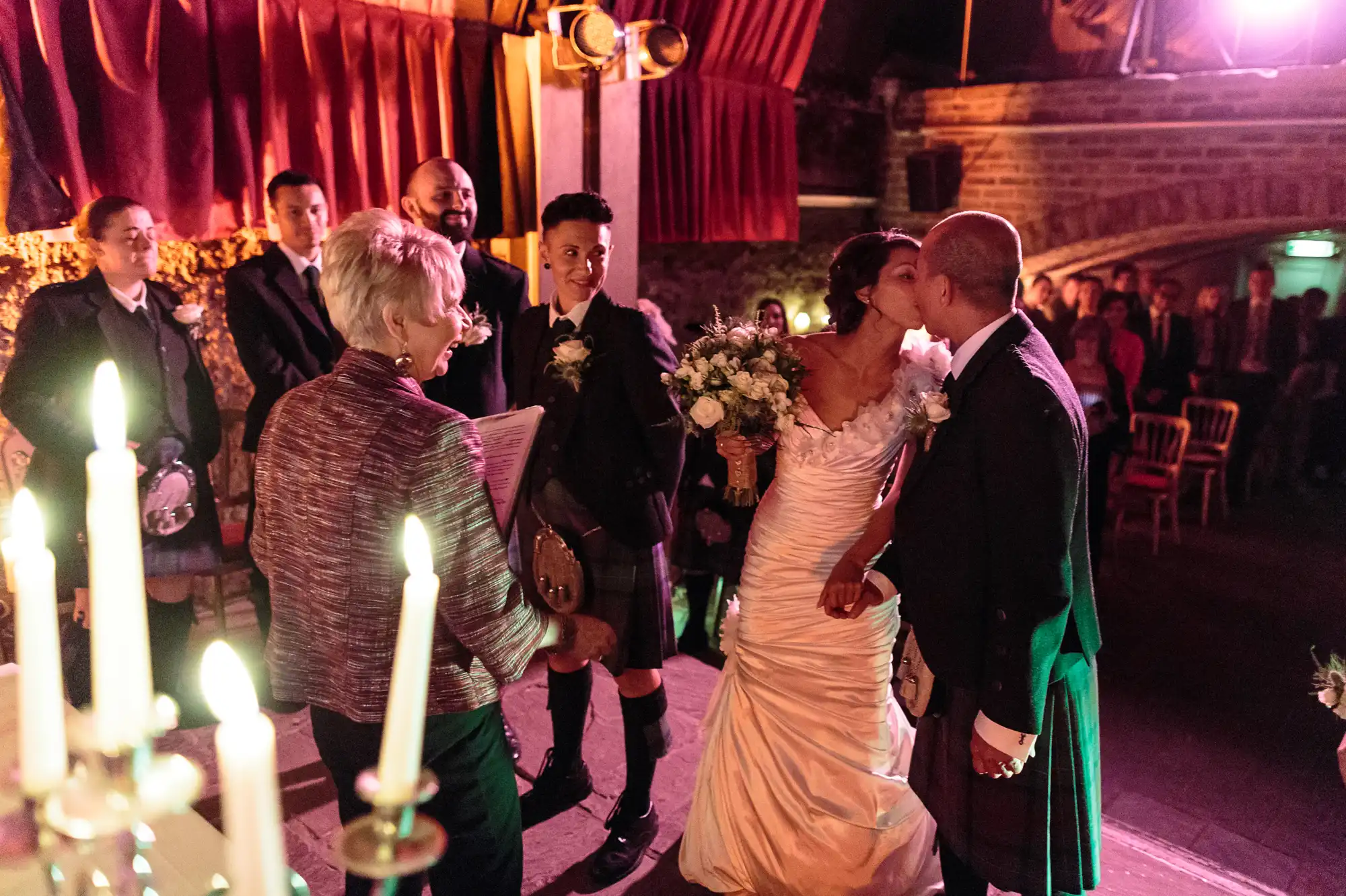A bride and groom kiss at their wedding ceremony in a warmly lit room with stone walls. The bride holds a bouquet, and several men in kilts stand nearby. An officiant reads from a book, and candles glow in the foreground. Guests watch from chairs in the background.
