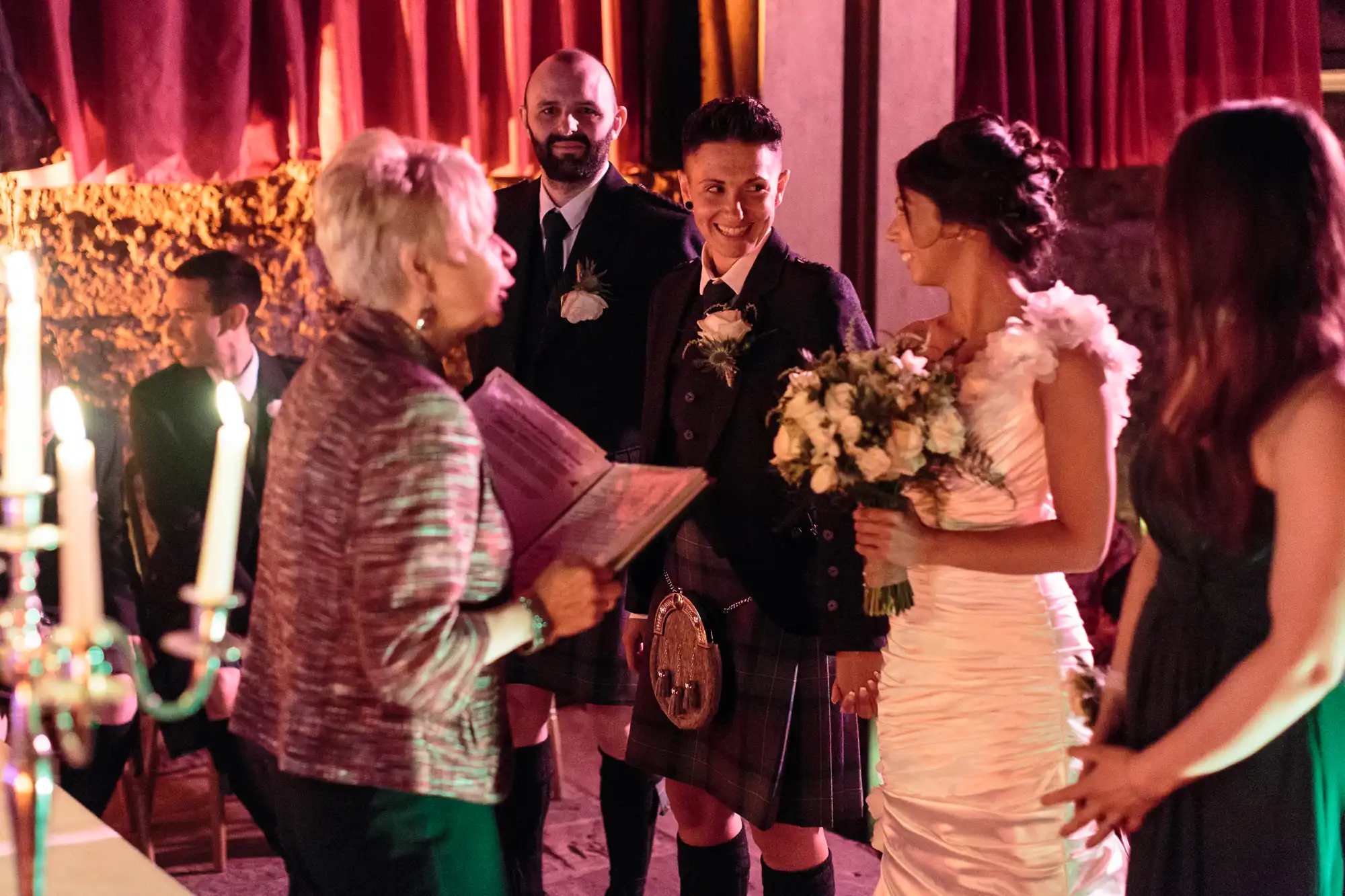 A wedding ceremony with two women, one in a kilt and the other in a white dress, smiling at each other. An officiant holds a book nearby. Two more people, one in a suit and another in a dress, stand beside them. Warm lighting and red curtains set the scene.