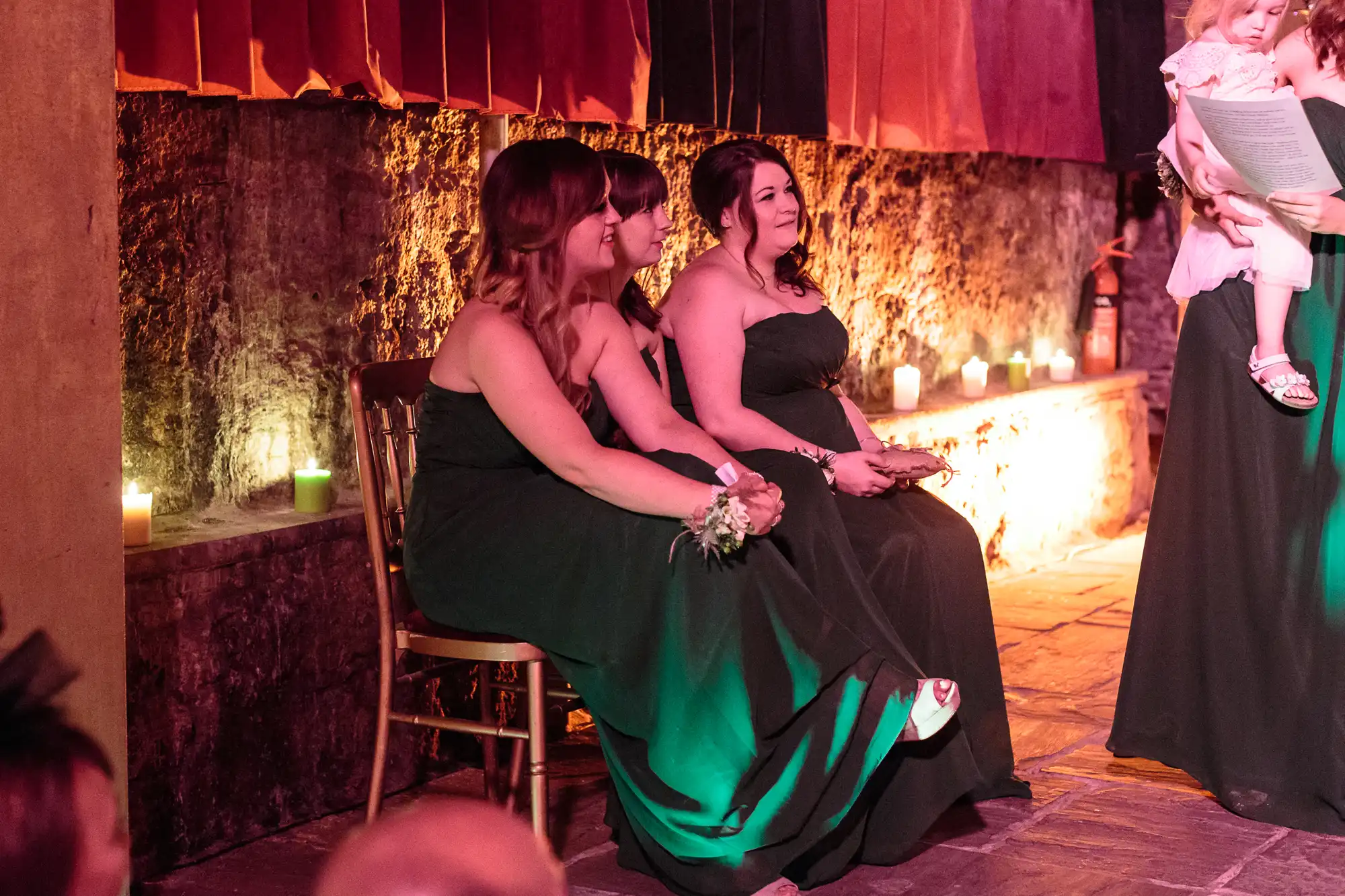Three women in dark green dresses sit on chairs at an indoor event with stone walls and candles. They are watching something off-camera, smiling, with hands in their laps. Warm lighting highlights their faces and the setting appears formal or festive.