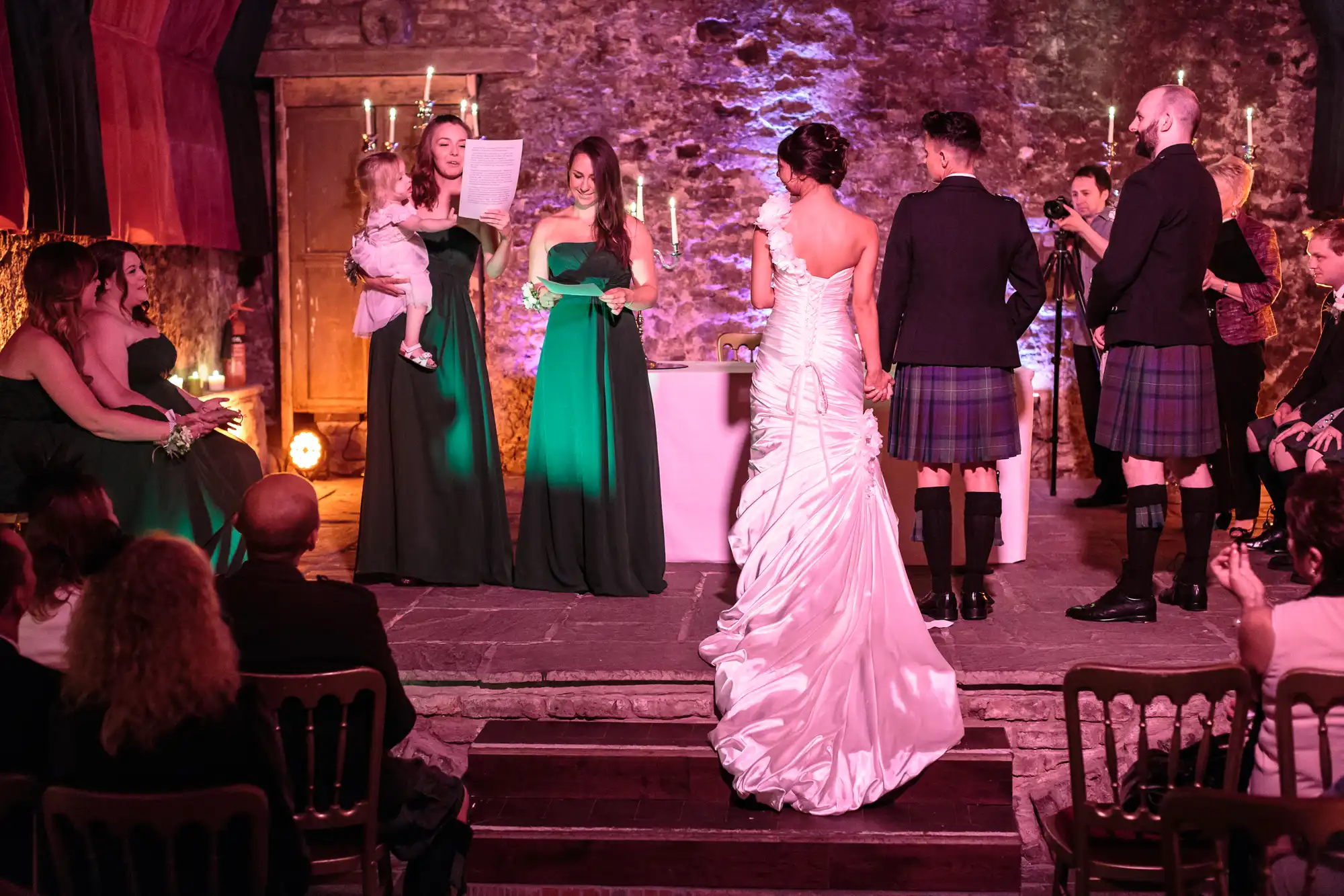 A bride in a white dress stands beside a groom in a kilt at an indoor wedding ceremony. Bridesmaids in green dresses, one holding a child, read from papers. Guests sit watching. Stone walls and candles create a warm, rustic atmosphere.