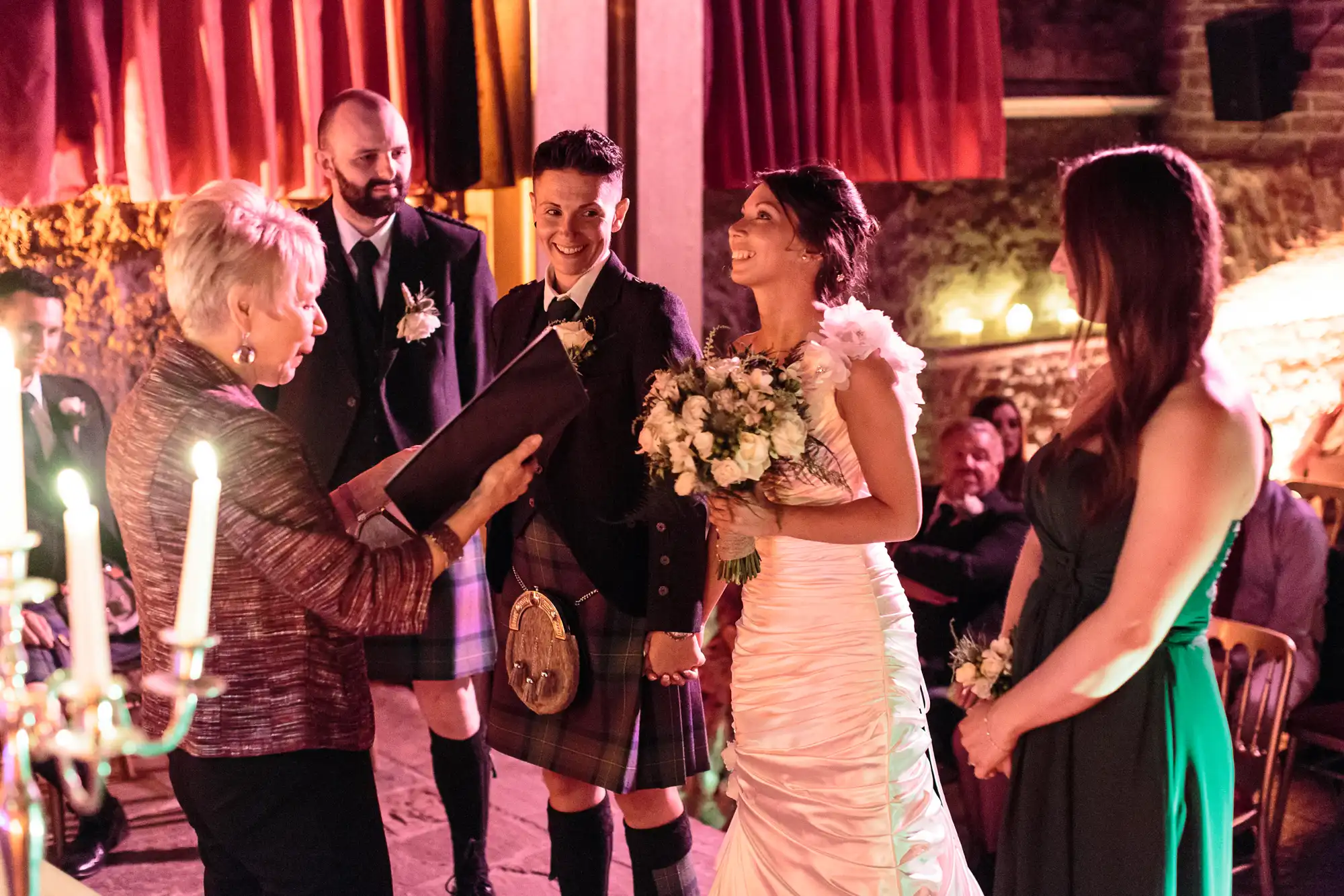 A wedding ceremony indoors shows a smiling bride in a white dress holding a bouquet, standing beside the groom in a kilt. An officiant reads from a book, whilst another woman in a green dress stands nearby. Warm lighting and red curtains create a cosy atmosphere.
