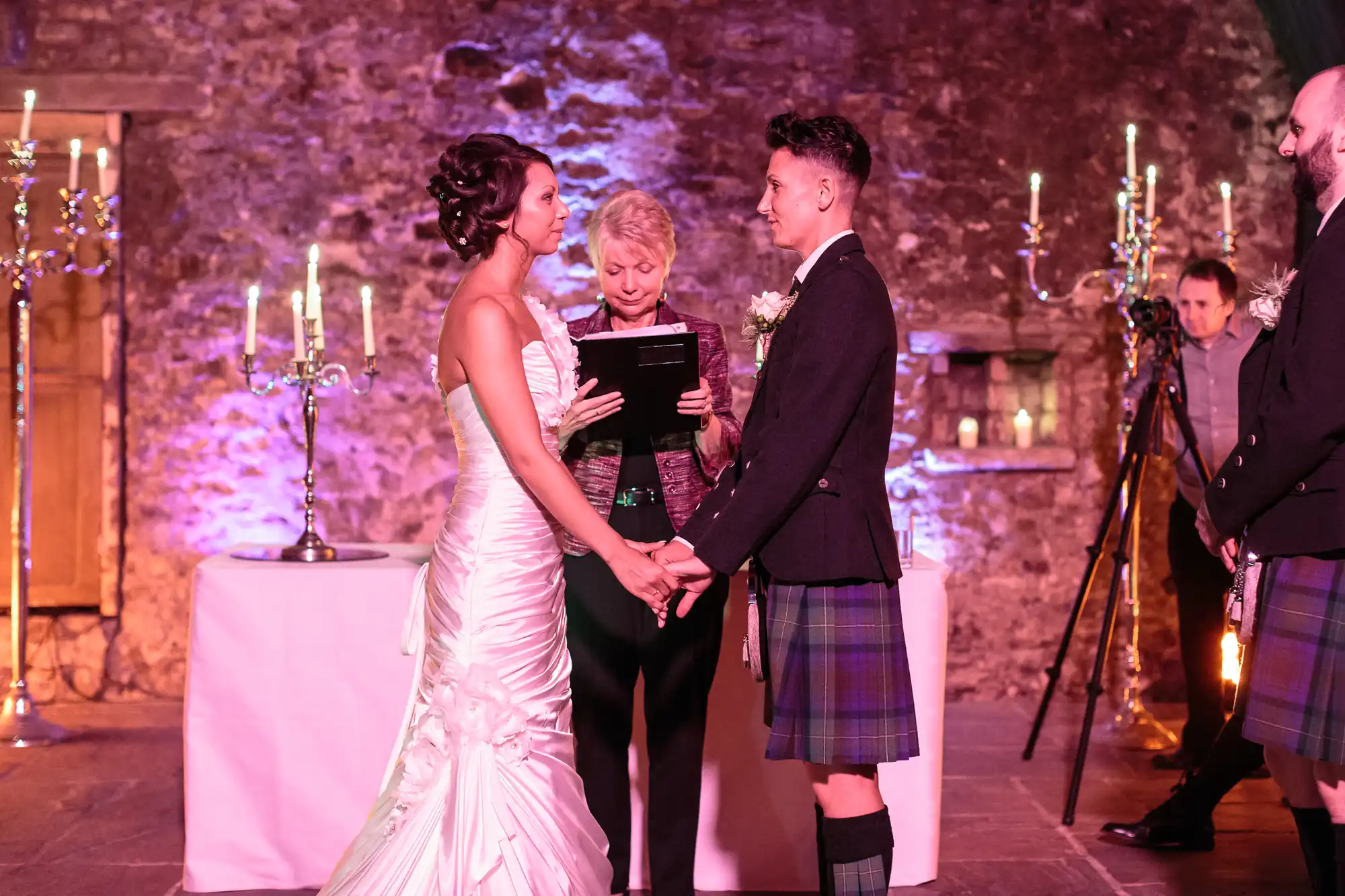 A bride in a white dress and a groom in a kilt hold hands, facing each other during a wedding ceremony. An officiant stands between them, reading from a book. The background features stone walls, candles, and soft purple lighting.