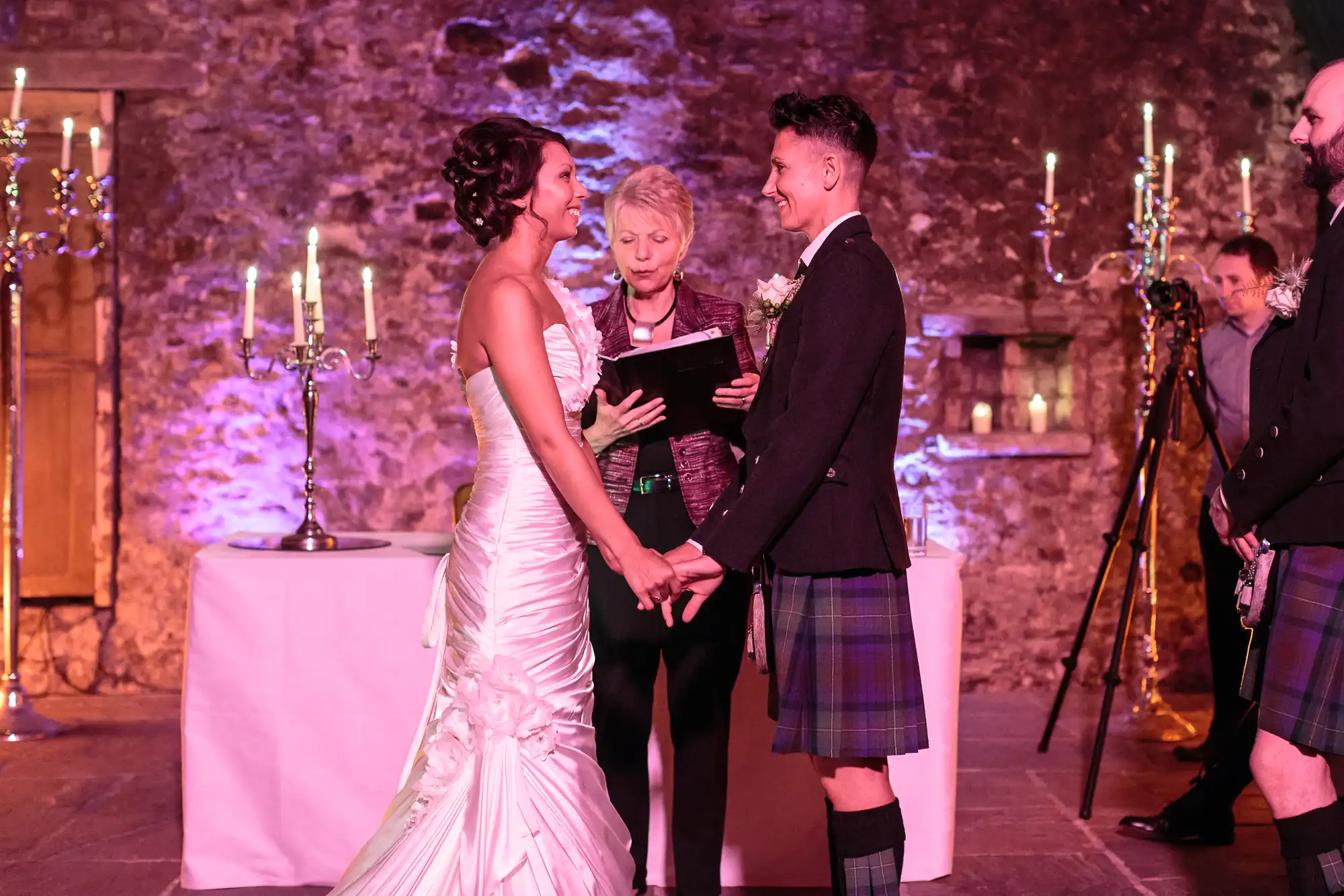 A bride and groom, both smiling and holding hands, stand facing each other during a wedding ceremony. The groom wears a kilt; the bride wears a white gown. An officiant stands behind them, with candelabra and a stone wall in the background.