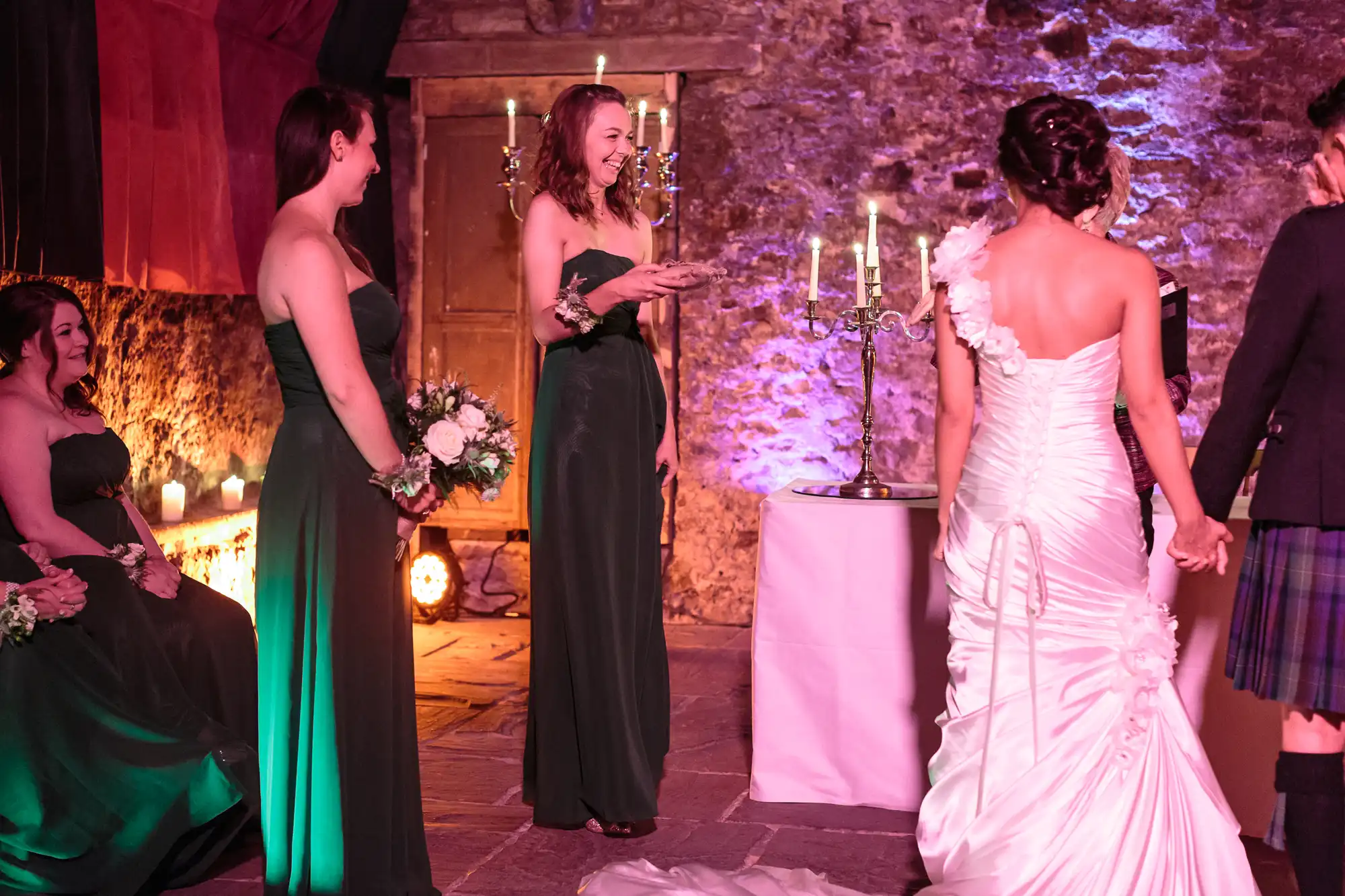 A bride in a white gown stands holding hands with a person in a kilt. Bridesmaids in green dresses smile and hold bouquets. The setting is indoors with stone walls, wooden doors, and lit candelabra, creating a warm, festive atmosphere.