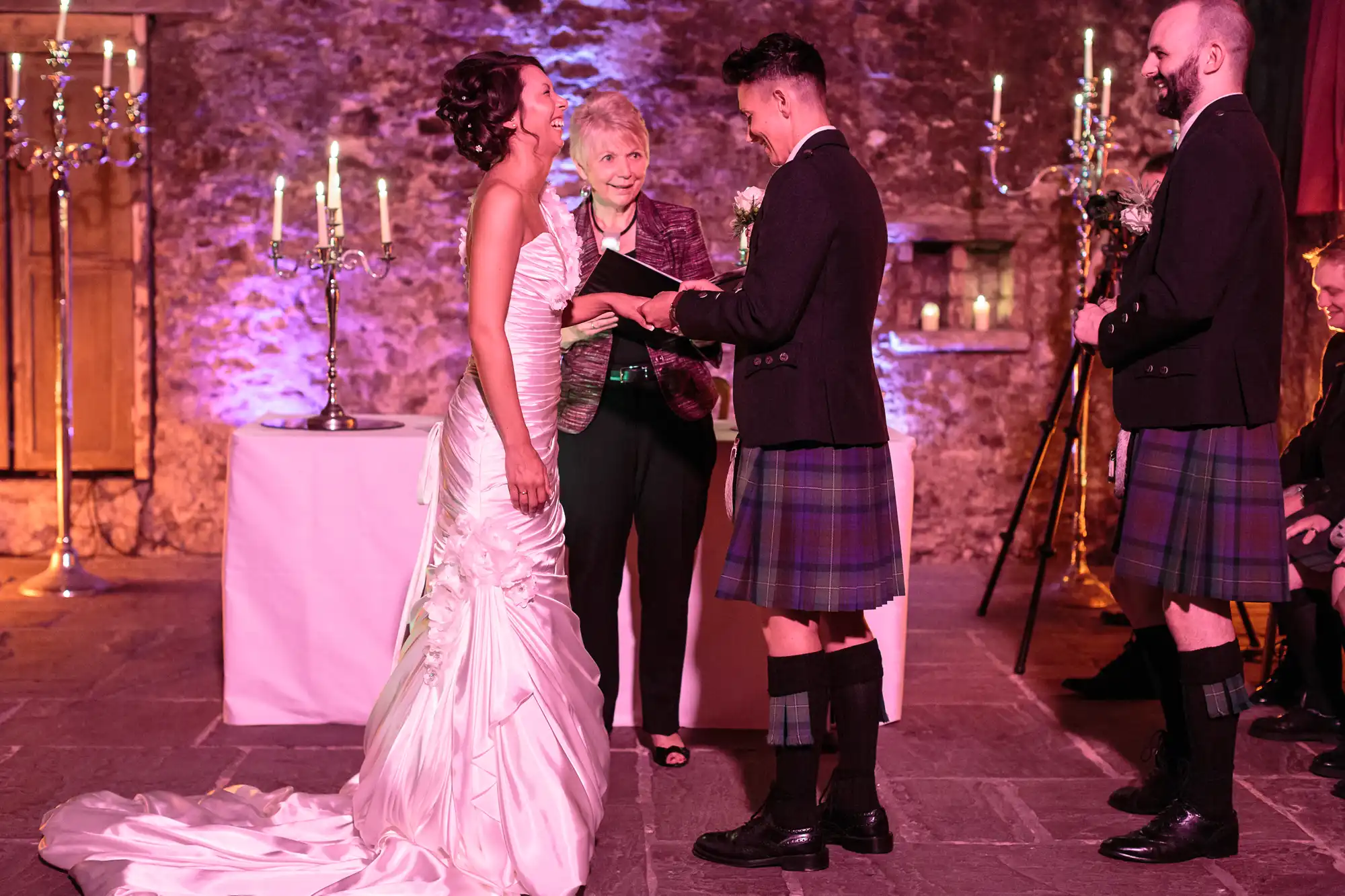 A bride in a white gown and a groom in a kilt exchange rings, smiling, during their wedding ceremony. An officiant stands between them, holding a book. Another man in a kilt stands to the side. The stone-walled venue is softly lit with candles.