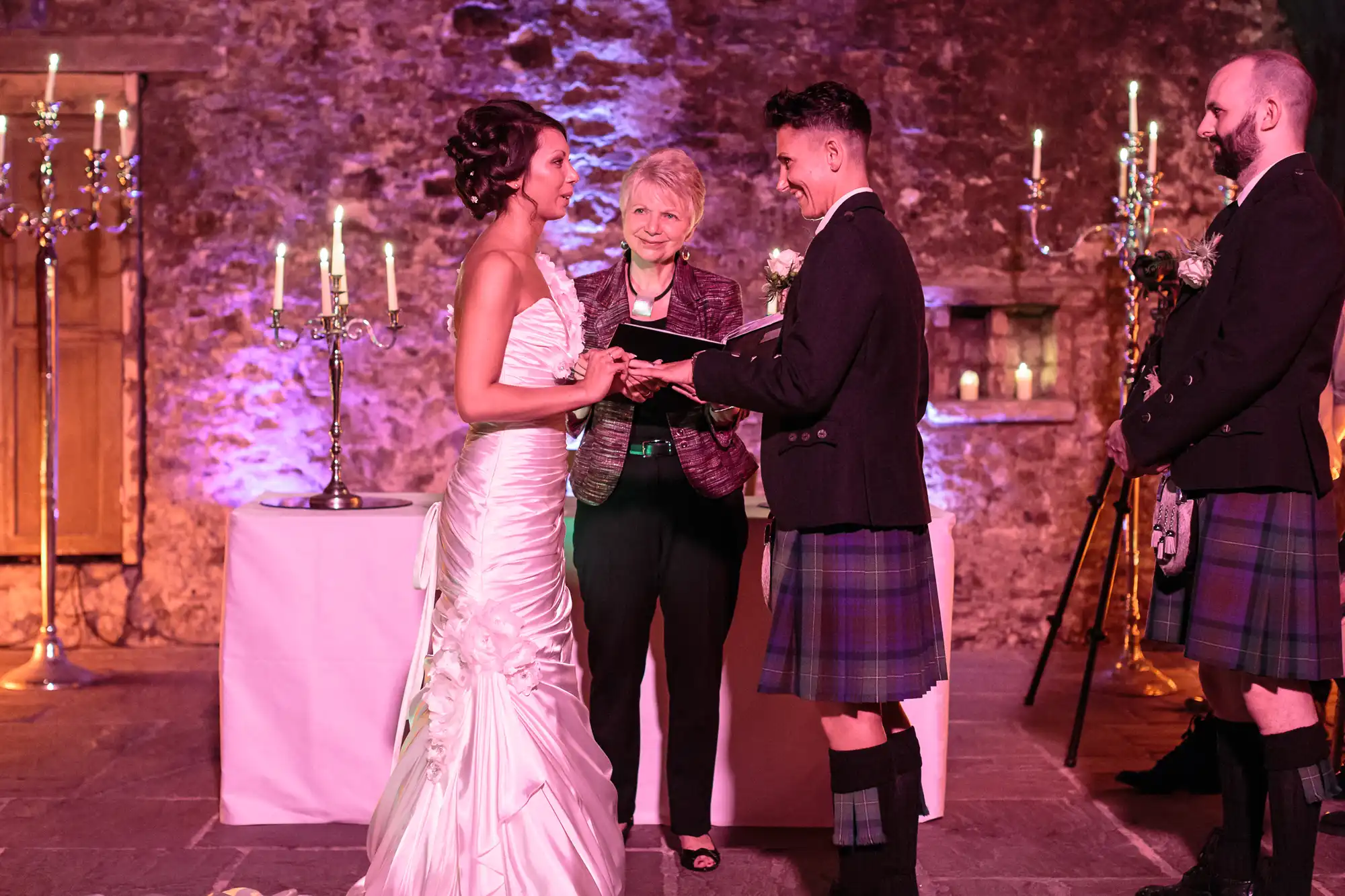 A bride in a white gown and a groom in a kilt exchange rings at an indoor ceremony. An officiant stands between them, smiling. Another man in a kilt stands to the groom’s right. The stone walls are lit with purple light and tall candelabras are behind them.