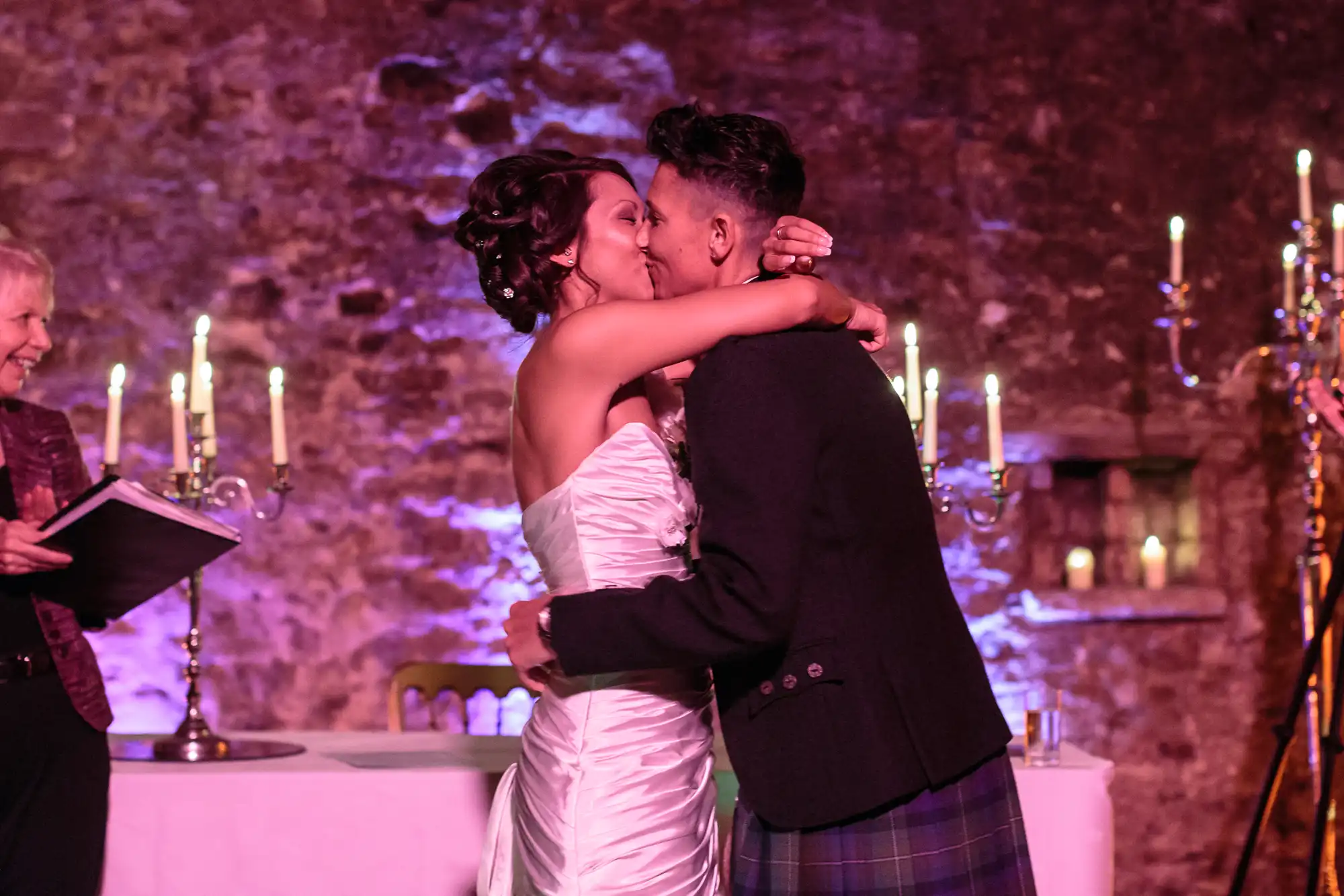 A bride in a white strapless gown hugs and kisses a groom in a dark jacket and tartan kilt. They are in front of a stone wall with lit candelabra. An officiant stands nearby, smiling and holding a book. The scene is warmly lit with purple accents.