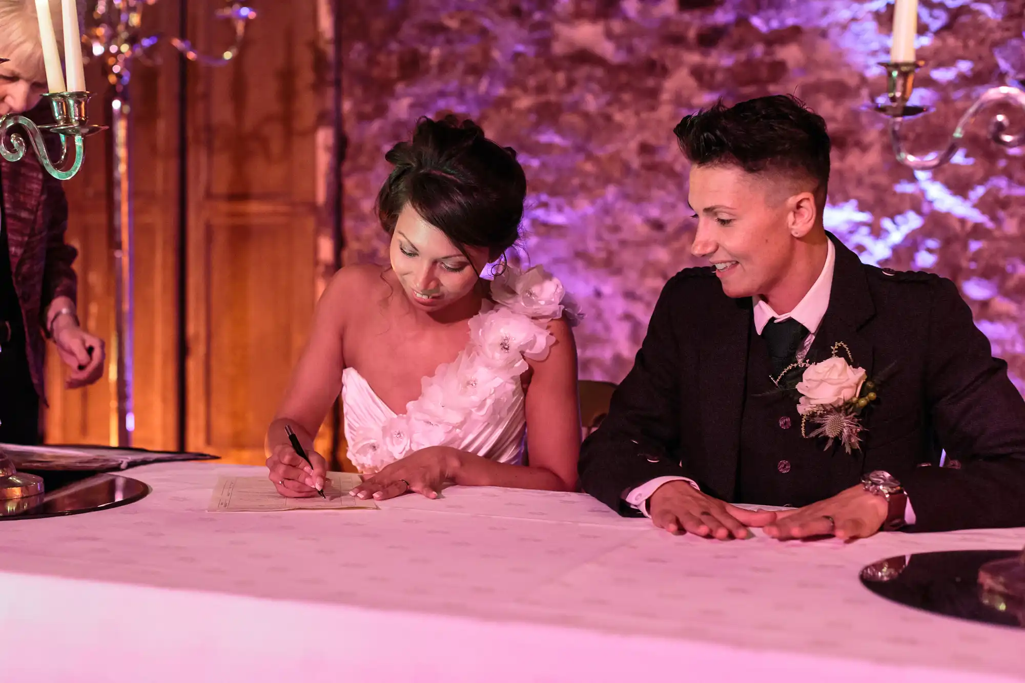 Two women at a wedding ceremony sit at a long, white-covered table. One, in a white dress with floral details, signs a document, while the other, in a black suit with a buttonhole, looks on smiling. The background features stone walls and warm lighting.