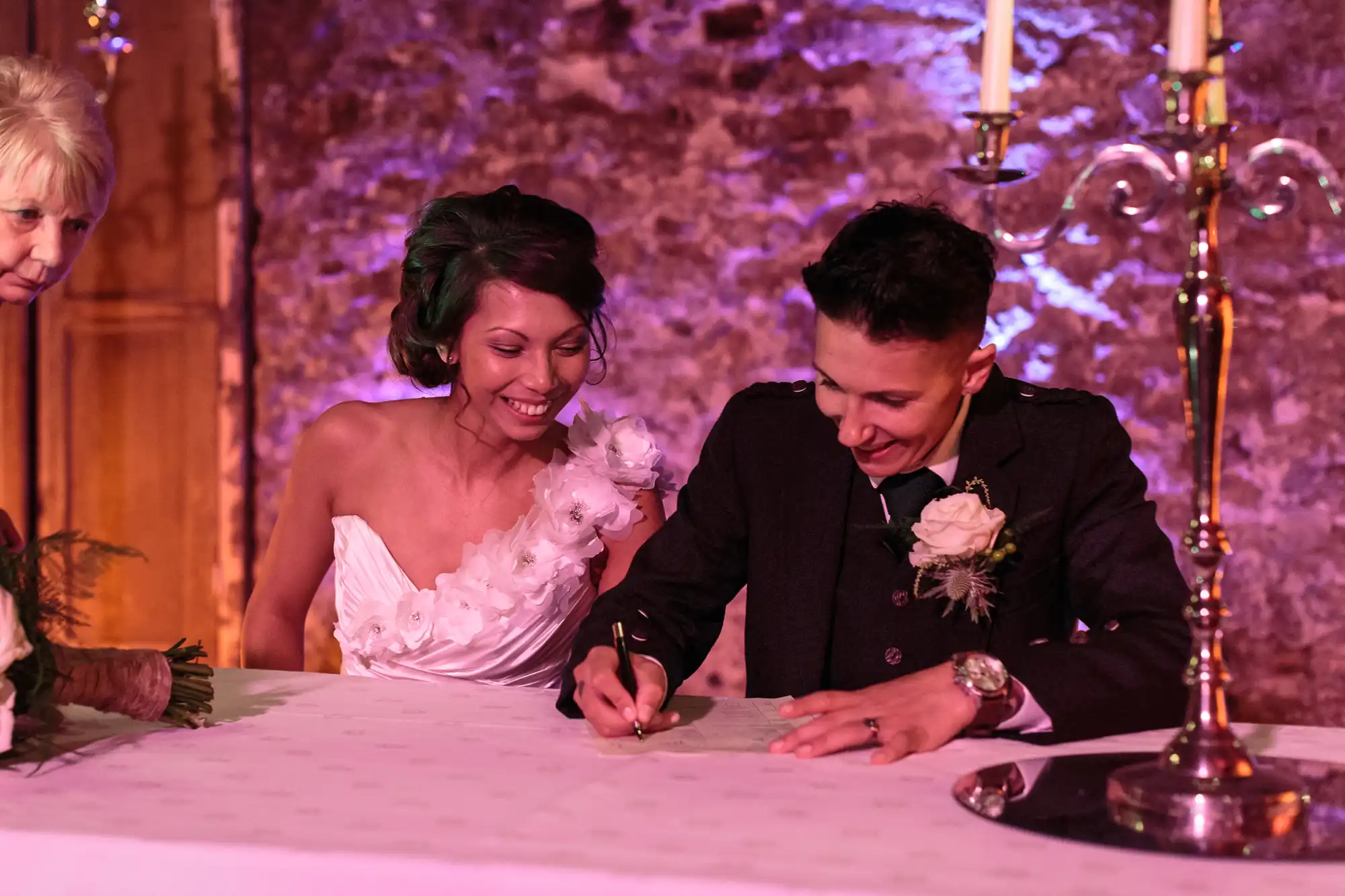 A bride in a white dress smiles whilst a groom in a dark suit signs a document on a table; a woman stands to the side. The setting is warmly lit, with a brick wall and a silver candelabrum in the background, suggesting a wedding ceremony.