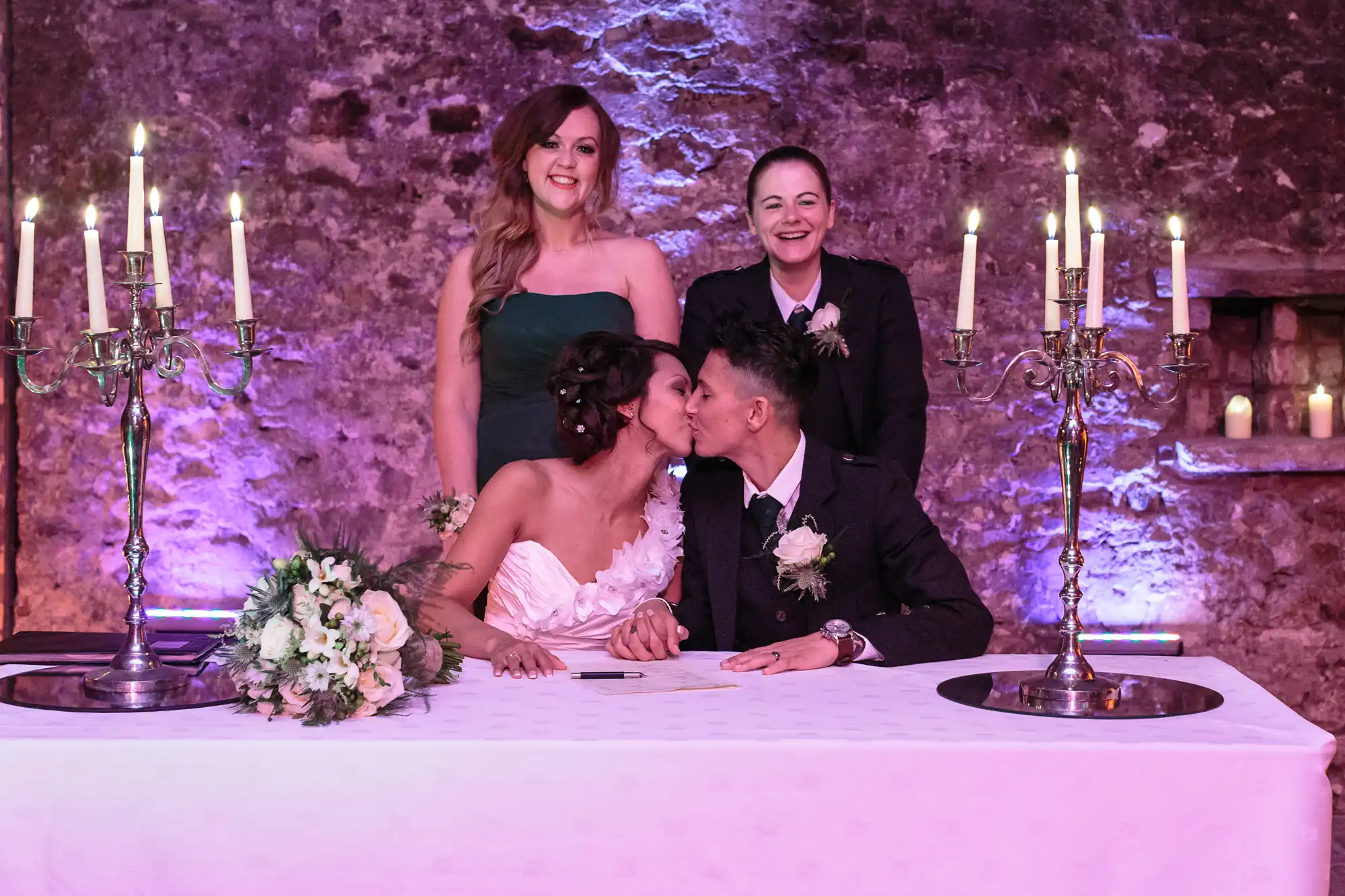 Two newlywed women in wedding attire sit at a table, kissing while holding hands. A bouquet and candles are on the table. Two women, one in a green dress and one in a suit, stand behind them smiling. The background is a rustic, purple-lit stone wall.