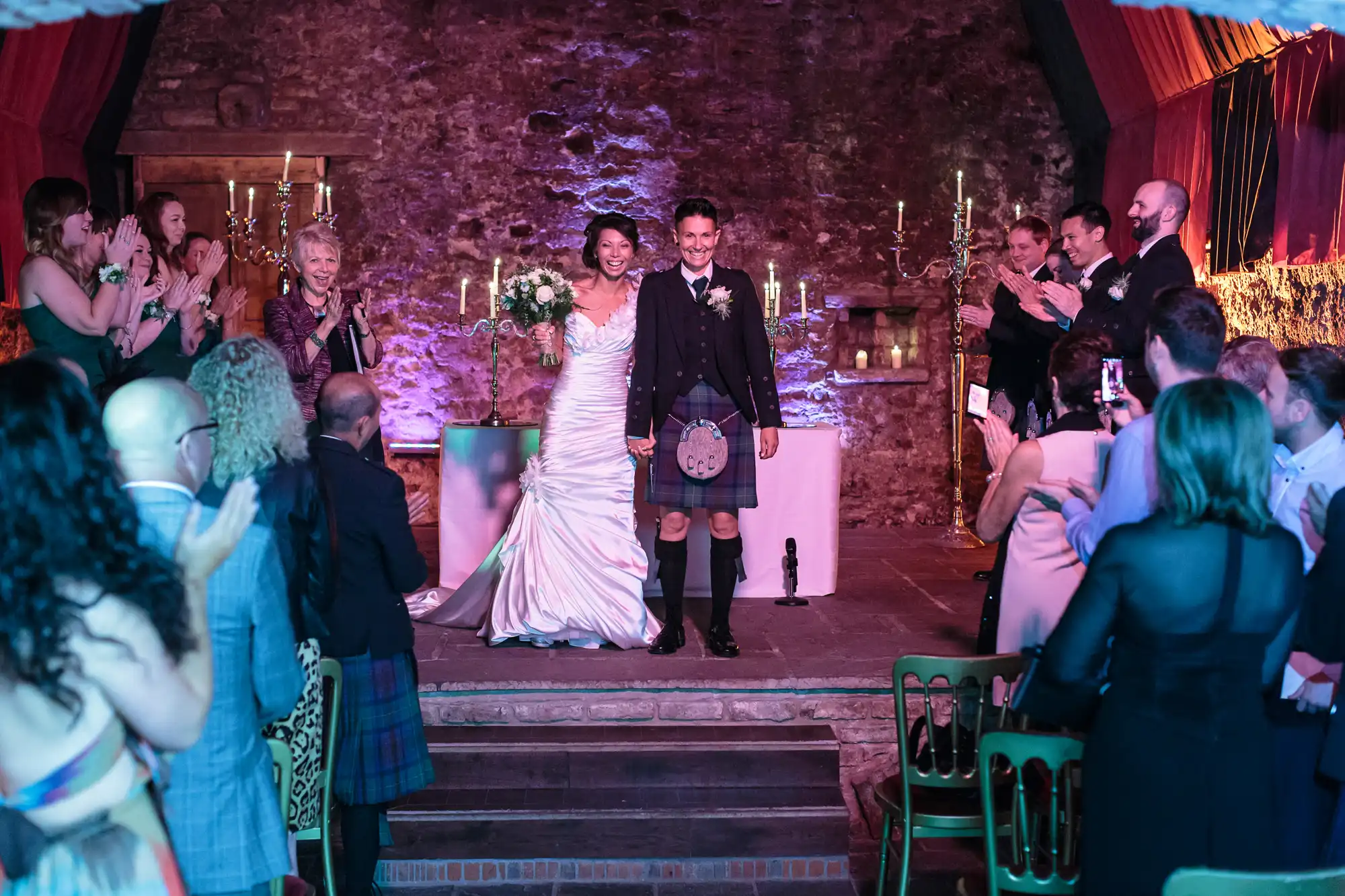 A newlywed couple stands smiling at the altar in a rustic stone venue, the bride in a white dress holding a bouquet and the groom in a kilt. Guests on both sides clap and cheer, with purple lighting and candles creating a festive, romantic atmosphere.