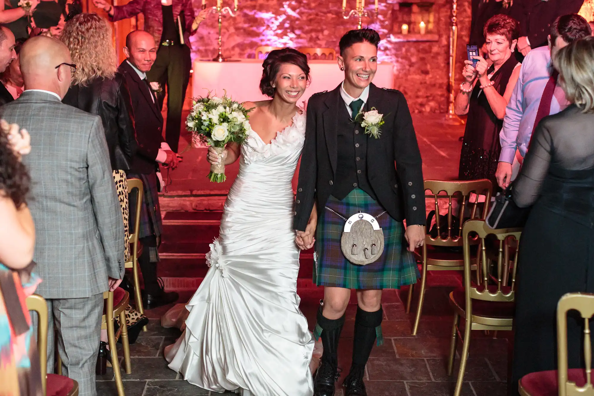 Two smiling brides walk hand-in-hand down an aisle. One wears a white dress holding a bouquet, the other a tartan kilt and jacket. Guests in formalwear sit and stand on either side, clapping and cheering in a warmly lit venue with brick walls.