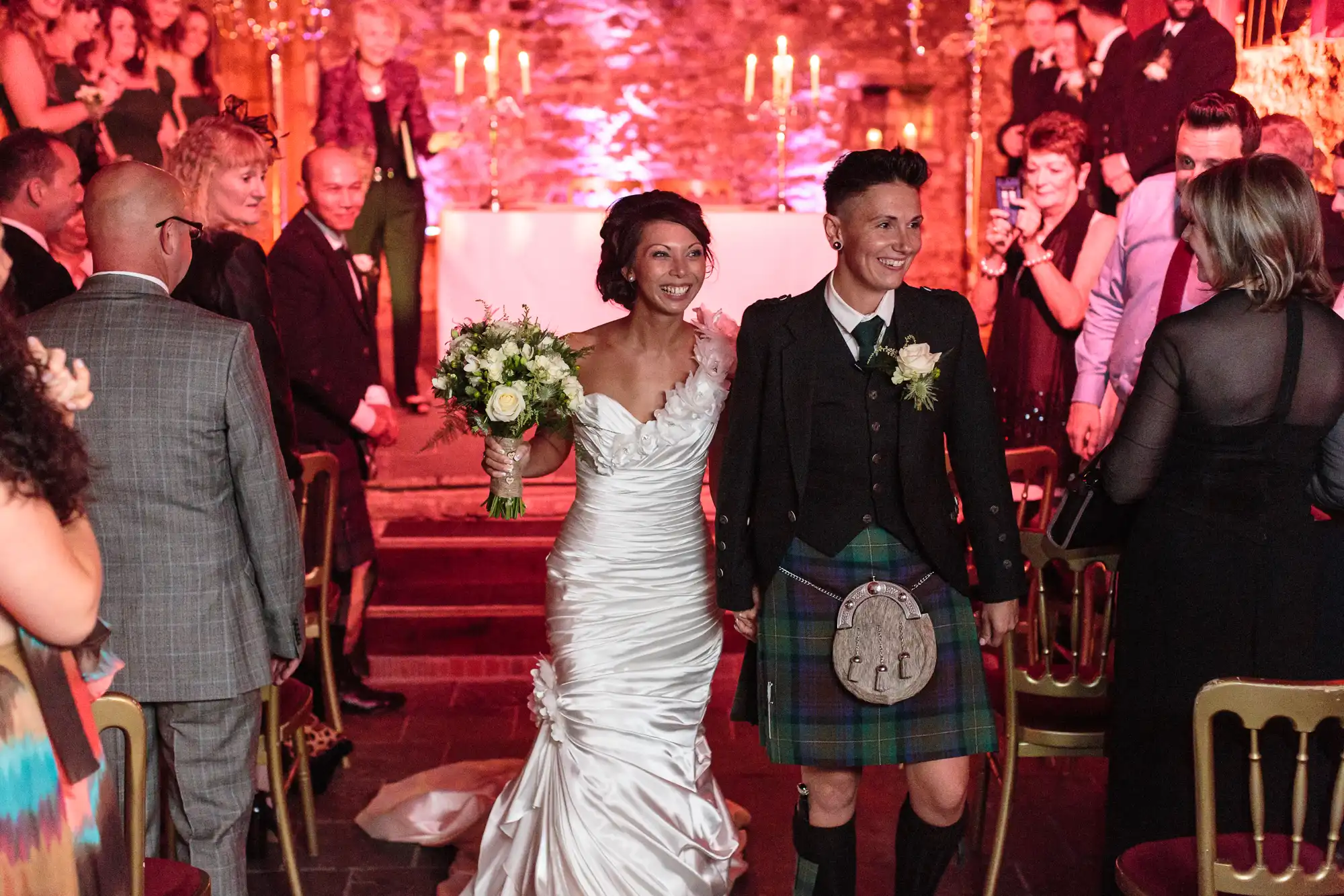 Two smiling brides walk down the aisle holding hands. One wears a white dress holding a bouquet; the other wears a kilt and jacket. Guests in formal attire watch and celebrate. The background is warmly lit with candles and rustic decor, creating a festive wedding atmosphere.