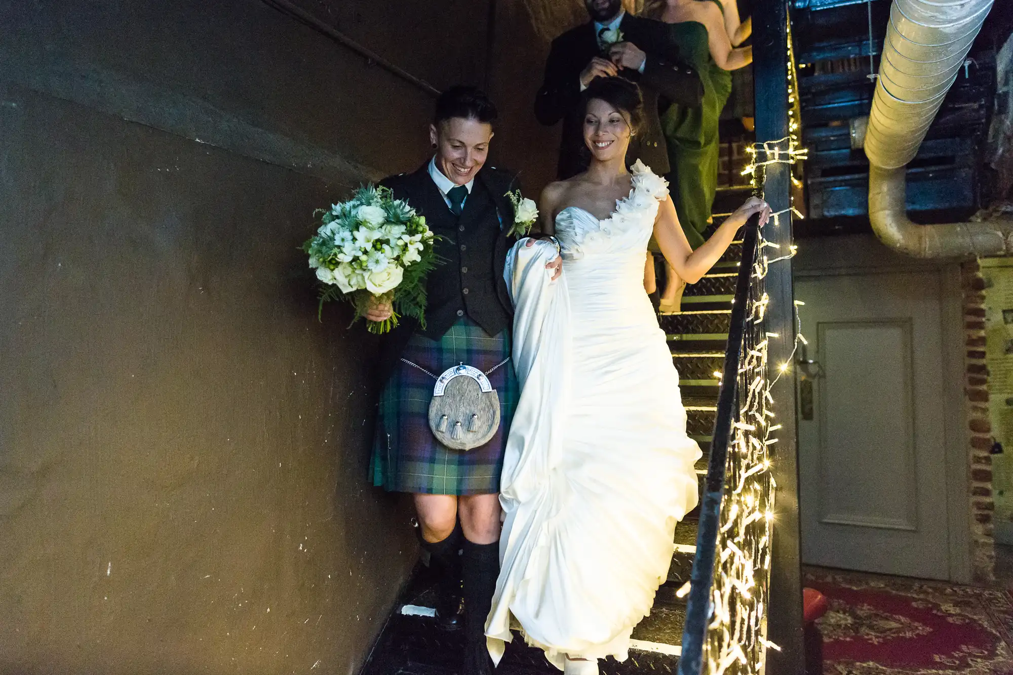 A smiling couple descends a staircase wrapped in fairy lights. One person wears a dark jacket and a checked kilt, holding a bouquet. The other wears a white, one-shoulder wedding dress, holding the dress. Both seem happy and joyful at the celebration.