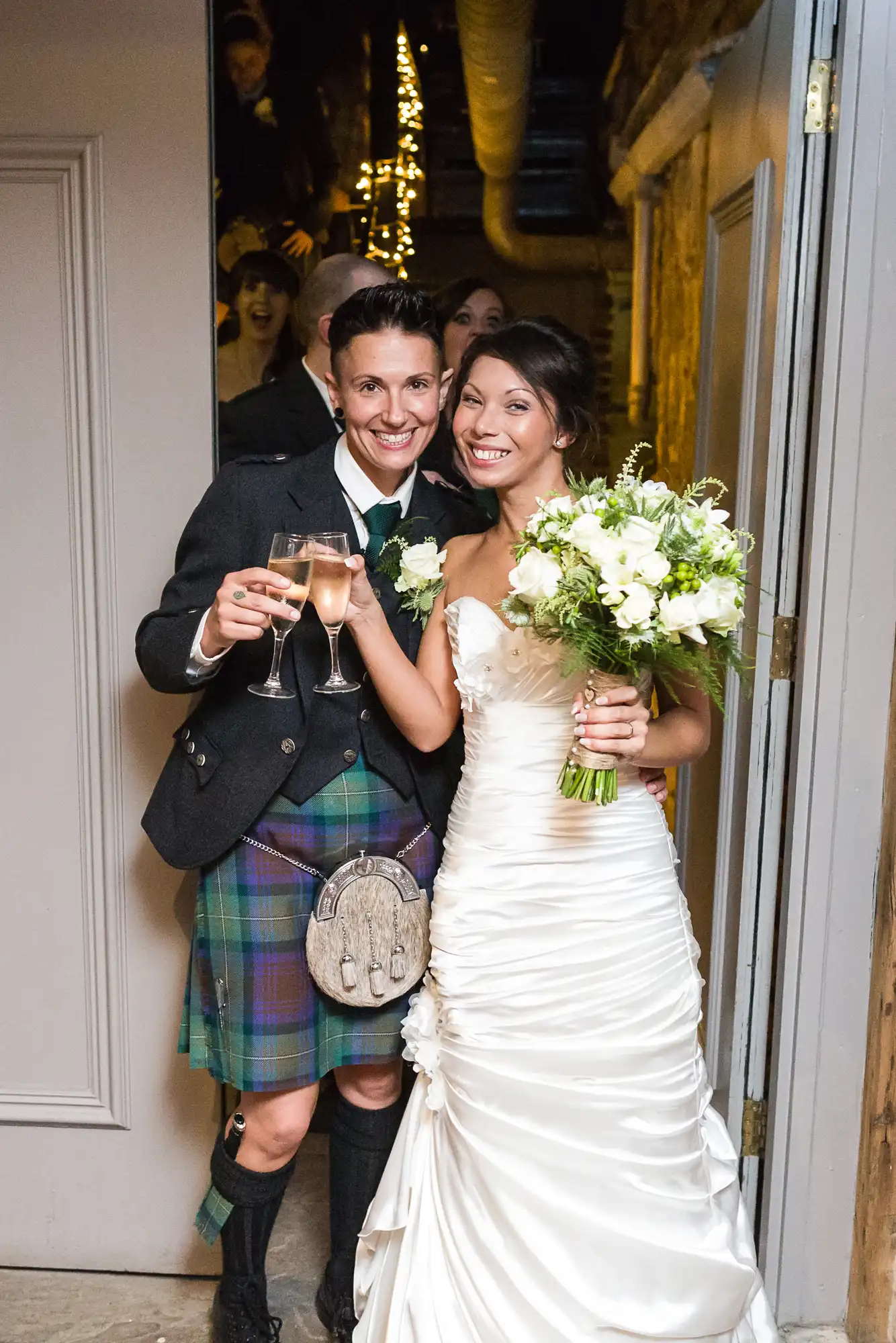 A smiling couple at their wedding, one in a checked kilt outfit holding a glass of champagne, the other in a strapless white wedding dress holding a bouquet and a glass of champagne. They stand in a doorway, with happy guests visible in the background.