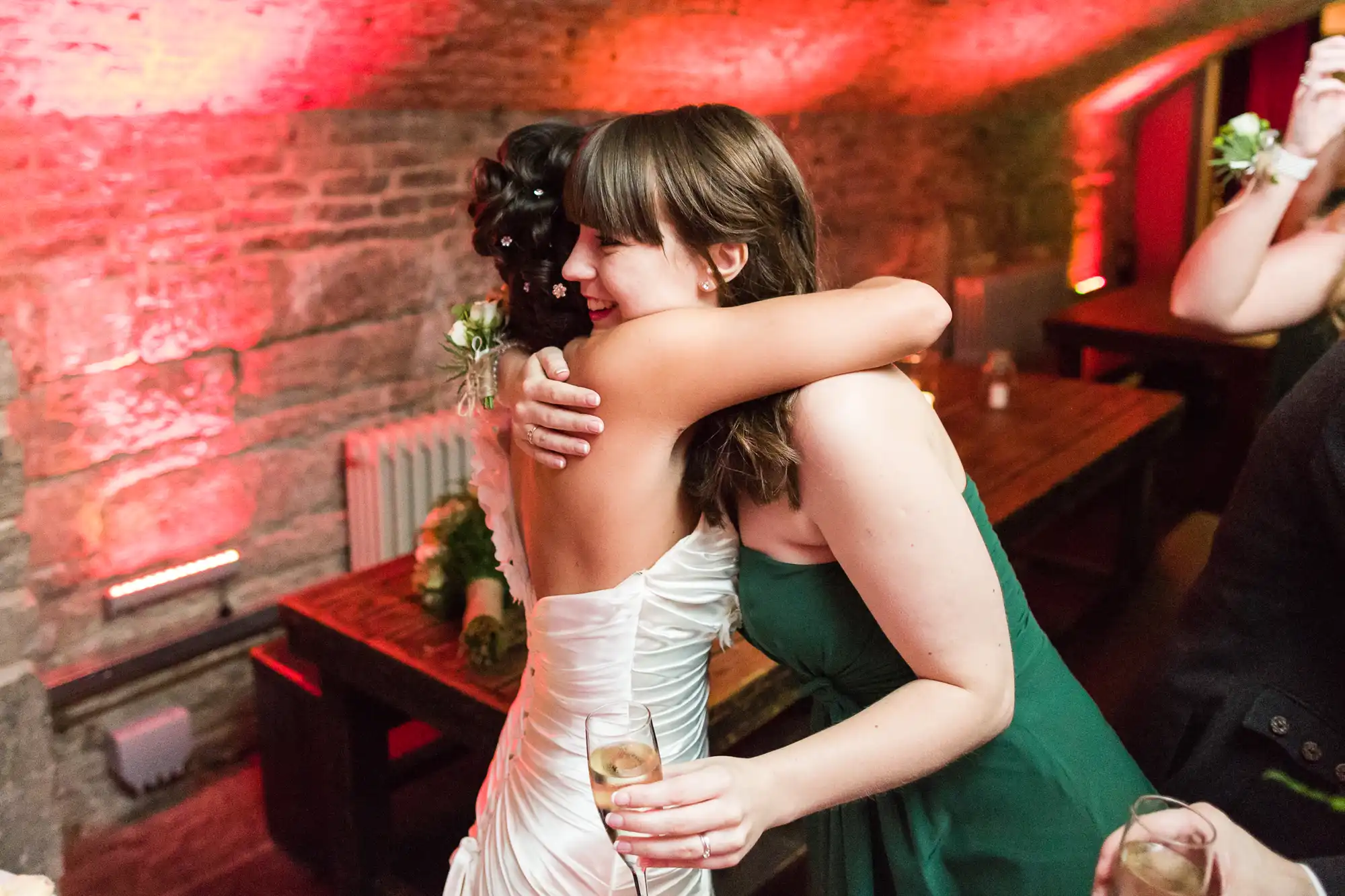 Two women hug warmly in a dimly lit room with exposed brick walls. One wears a white dress and holds a champagne glass; the other wears a green dress. Pink lighting adds a festive glow to the celebratory setting.