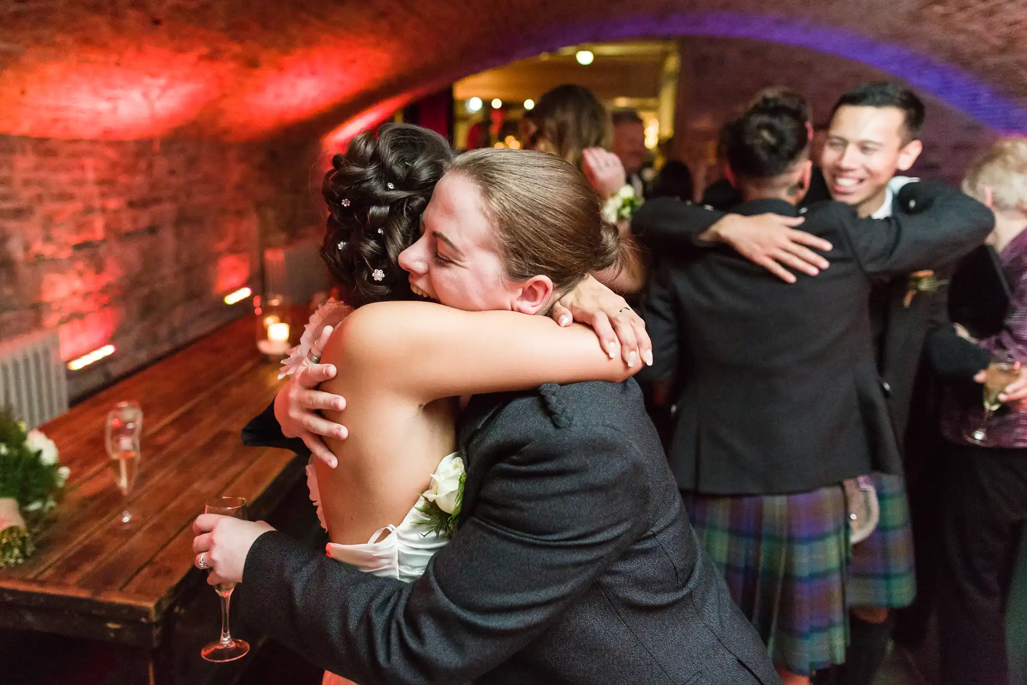 Two people embrace joyfully at a warmly lit celebration under a brick archway. Others in the background hug and smile, some in formal attire and kilts. A wooden table with candles and glasses adds to the festive atmosphere.