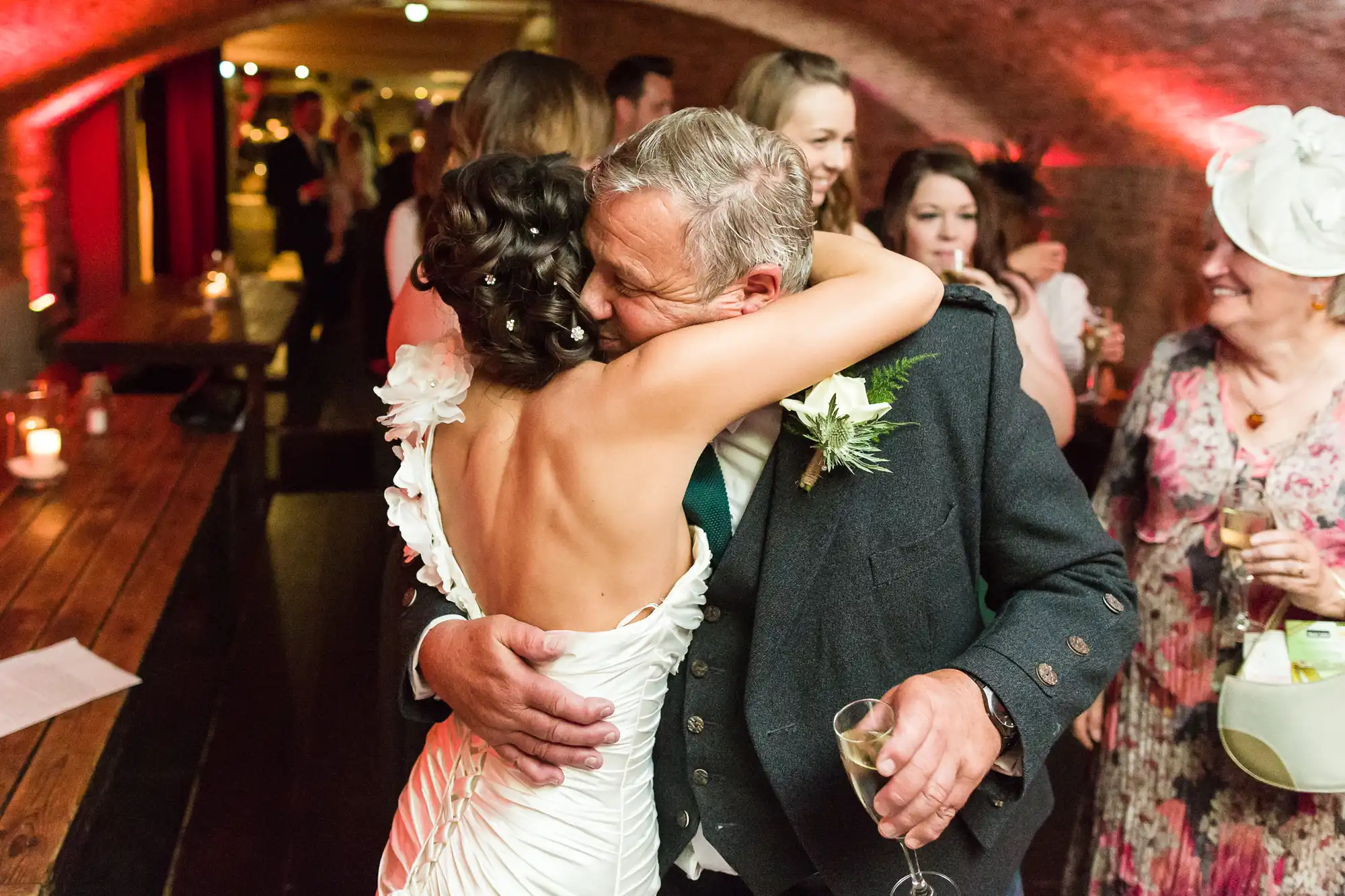 A bride in a white dress hugs an older man in a suit at a warmly lit wedding reception. Guests in festive attire smile in the background. The setting features rustic brick arches, wooden tables, and soft candlelight, creating an intimate, joyful atmosphere.